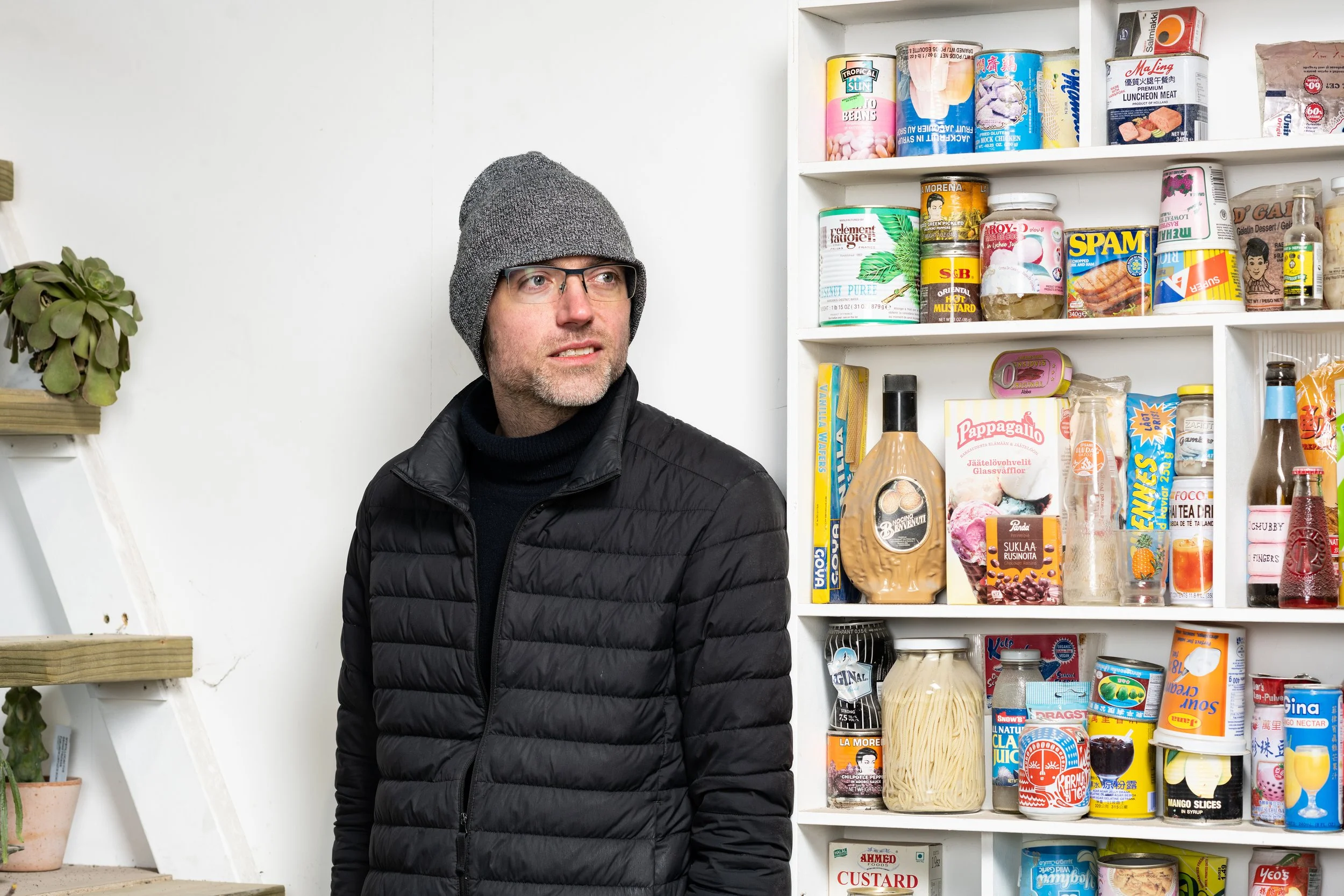 A man wearing glasses, a gray knit beanie, and a black puffer jacket standing in front of a white shelf filled with canned goods, jars, and packaged foods in a pantry or grocery store.