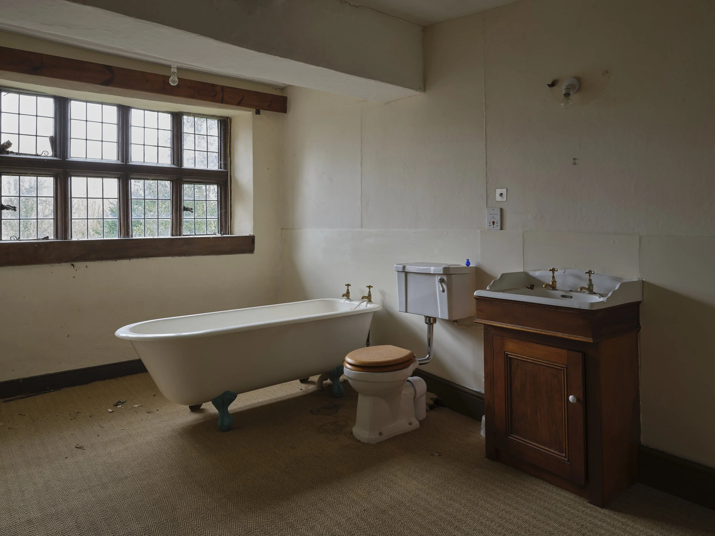 An old bathroom with a large window, a freestanding clawfoot bathtub with blue feet, a white toilet with a brown wooden seat, a porcelain sink with brass fixtures, a wooden cabinet under the sink, and an exposed light bulb on the ceiling. The walls a