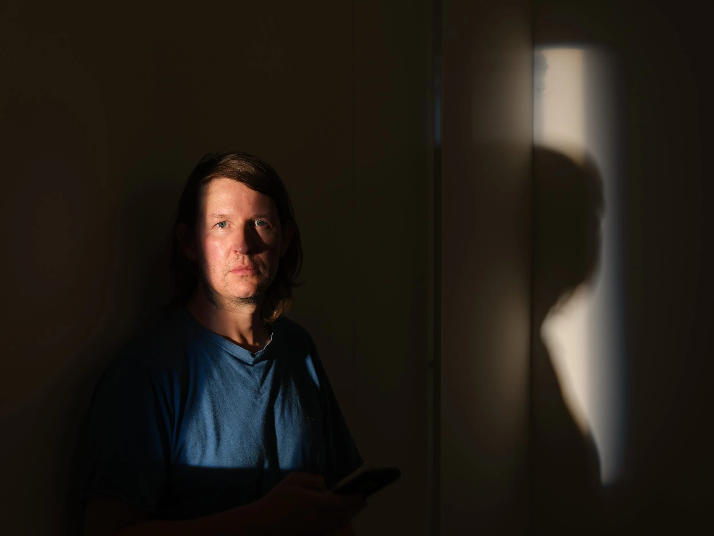 A man with shoulder-length brown hair, wearing a blue shirt and black jacket, stands in a dimly lit room, holding a smartphone. There is a bright light source creating a circular reflection on the wall behind him.