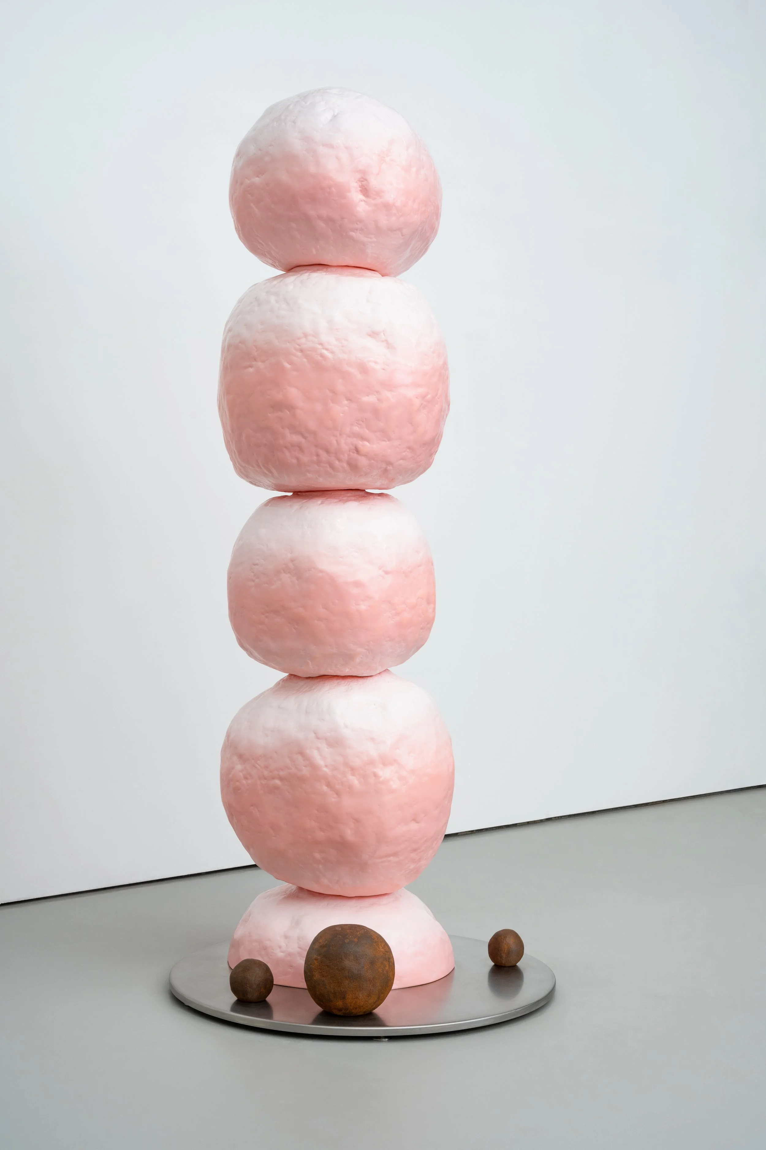 A tall sculpture of stacked pink and white rounded shapes resembling stones or candy, placed on a circular gray base, with several smaller brown spherical objects at the base, against a plain white wall background.
