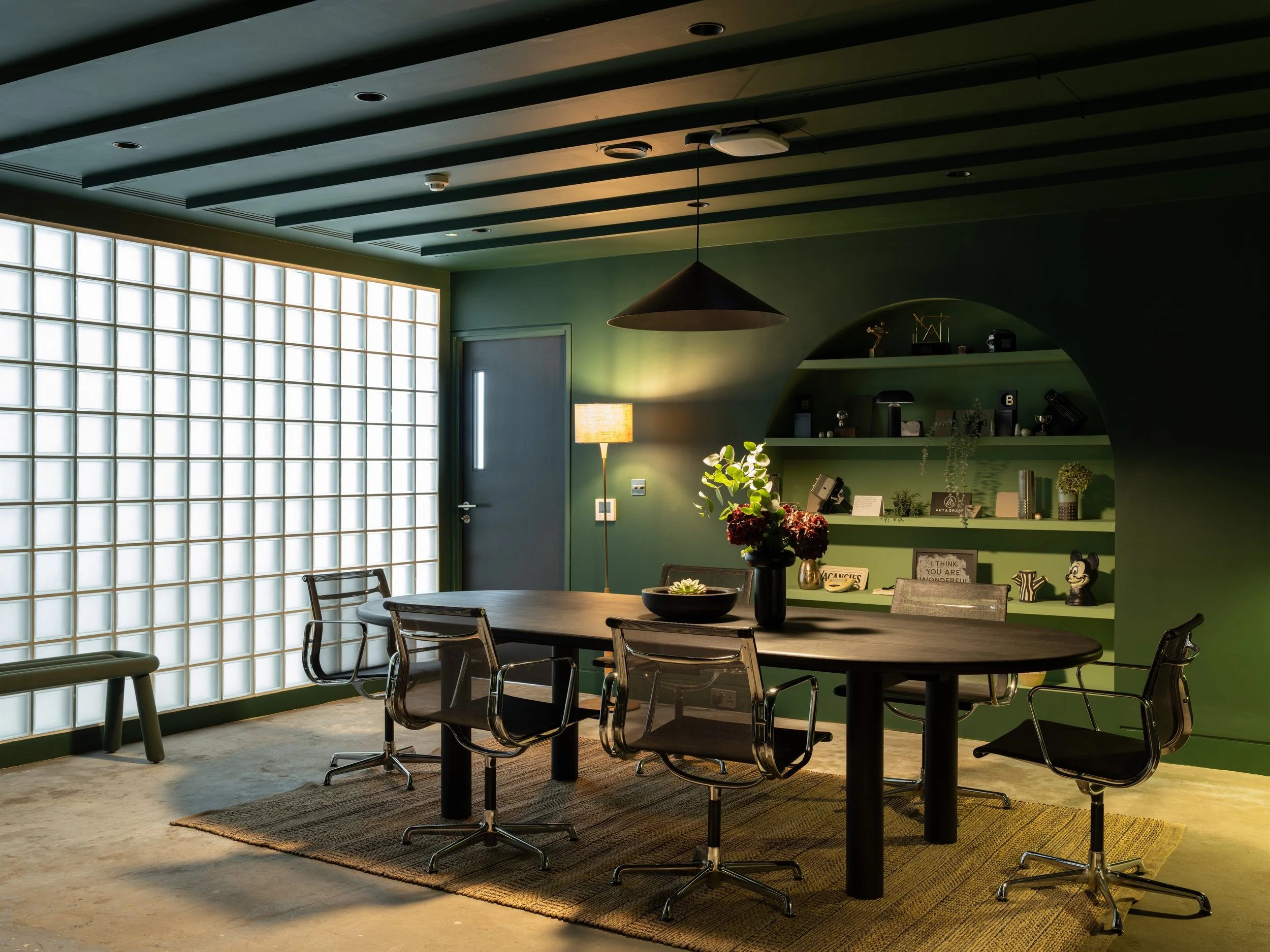 A modern dining room with a wooden table, six chairs with armrests, a green wall, and a large frosted glass window. There is a pendant lamp hanging above the table, a standing lamp, decorative shelves with plants and ornaments, and a Mickey Mouse fig