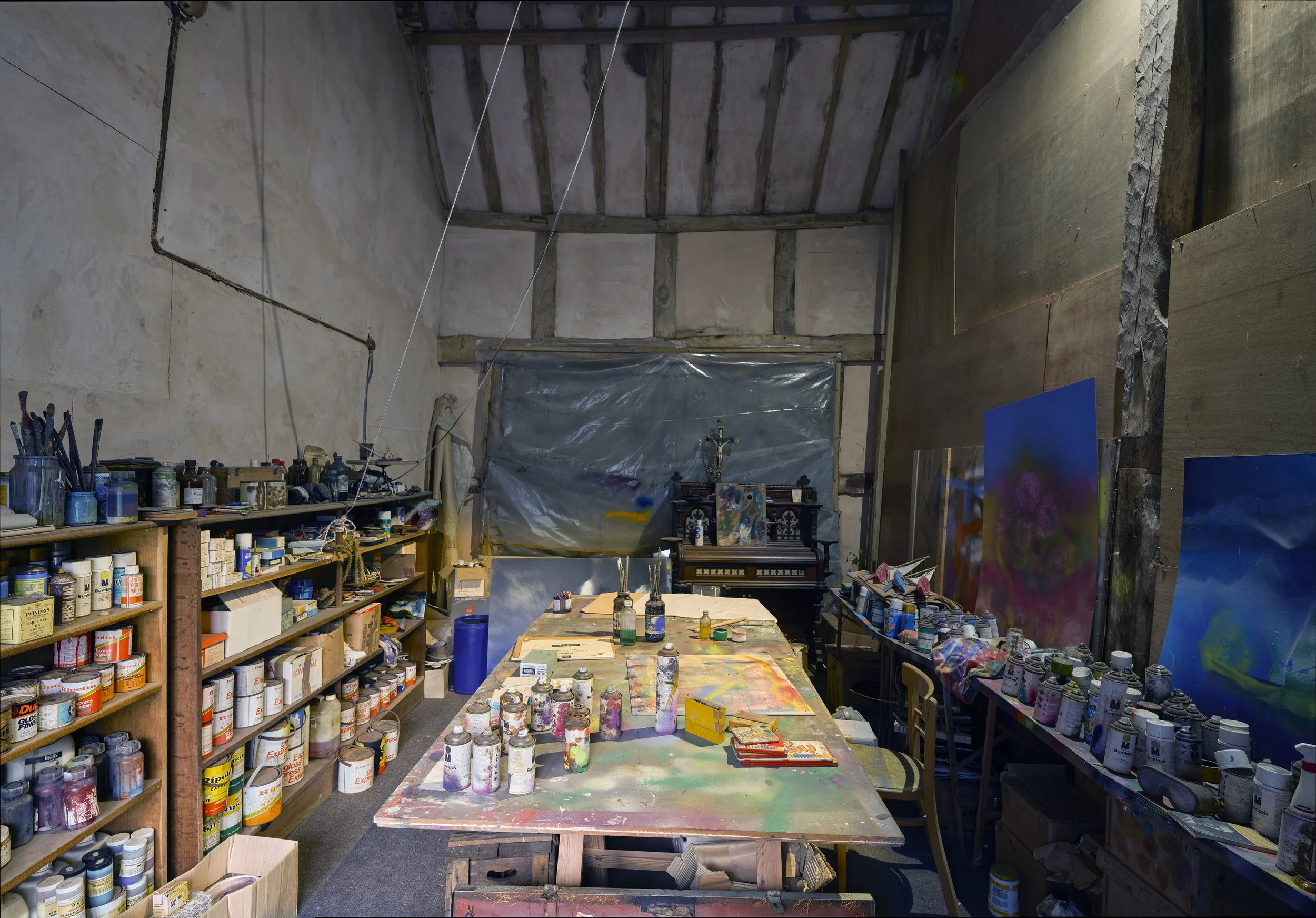 An artist's workshop with shelves filled with paint cans, brushes, and art supplies, and a table covered with colorful paints and paintings in progress.