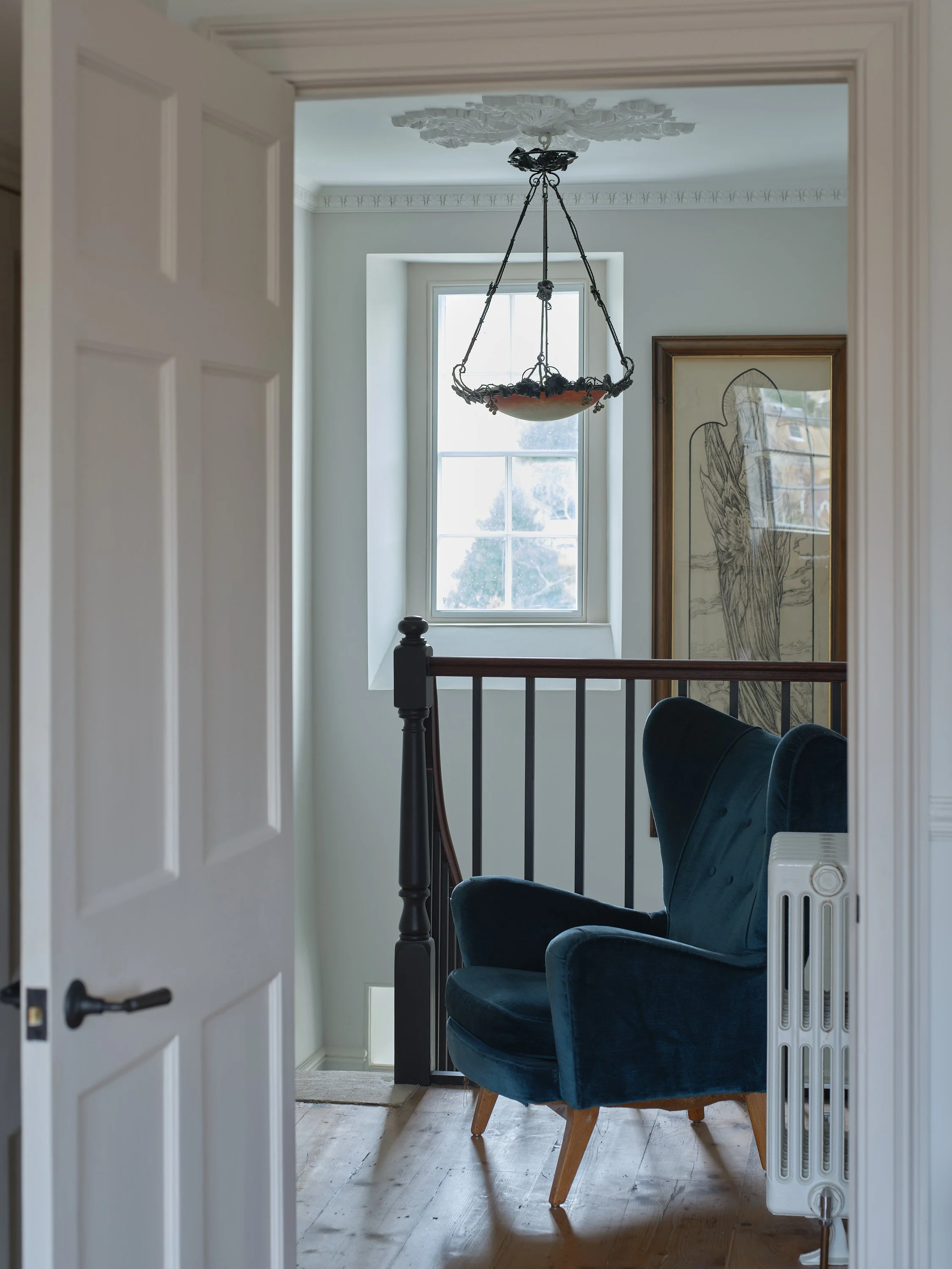 A door opens to a small sitting area with a blue upholstered armchair, a window, a framed drawing of a bird, and a hanging vintage-style chandelier.