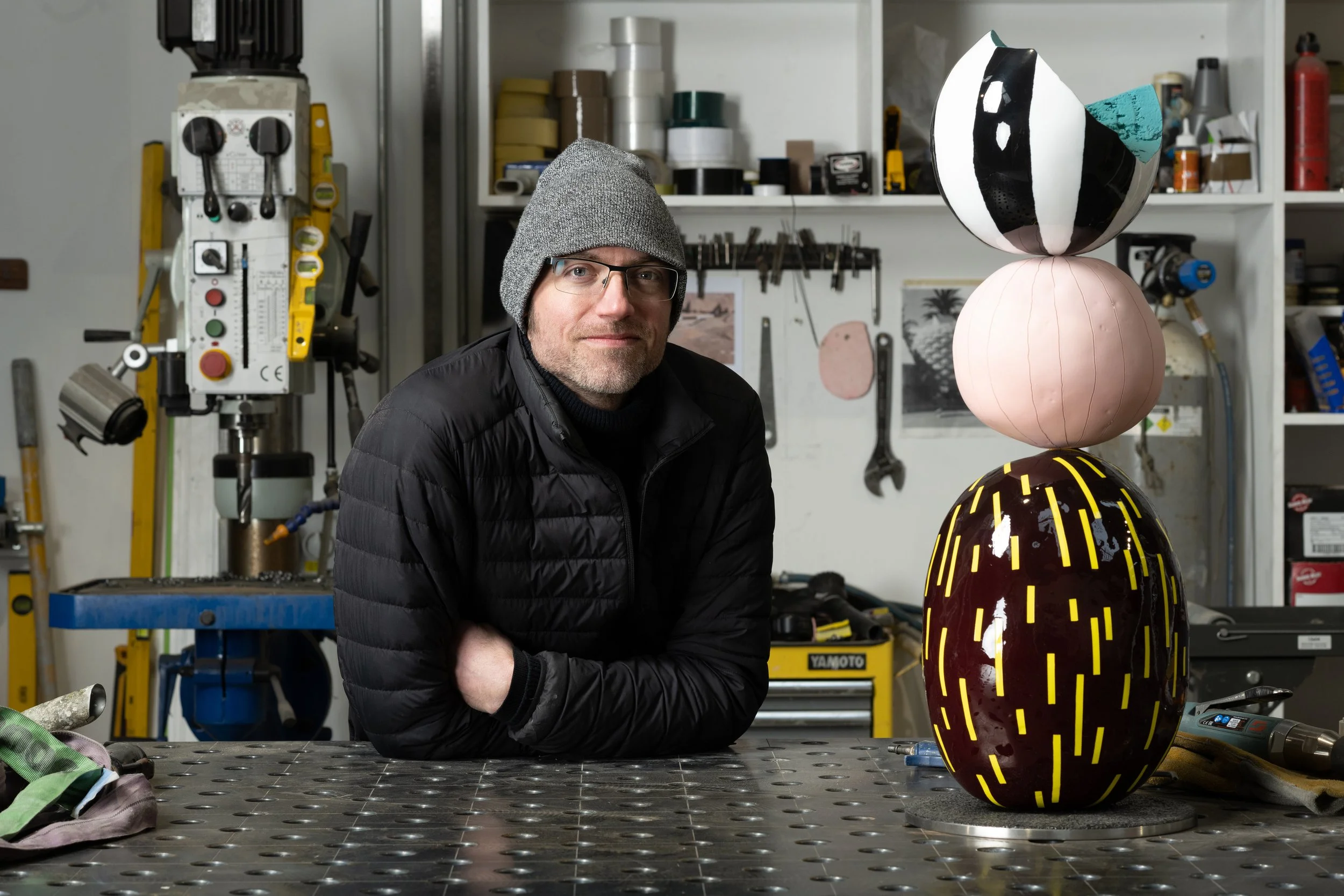 A man wearing glasses, a gray beanie, and a black jacket sitting at a workbench in a workshop with colorful, artistic sculptures in front of him. The workshop has tools and supplies on shelves in the background.