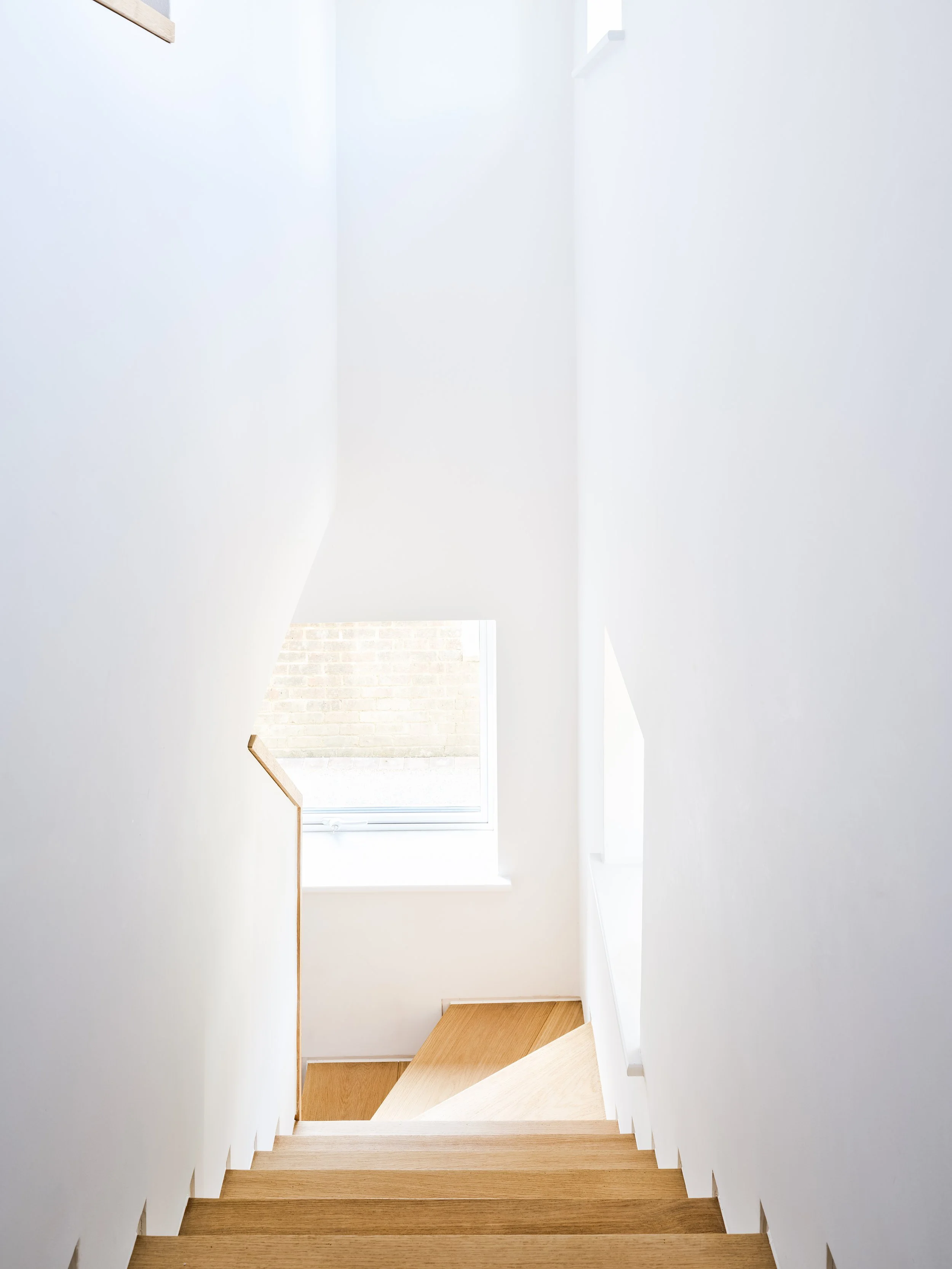 A staircase with wooden steps and white walls, leading to a door with a window that shows a brick wall outside.