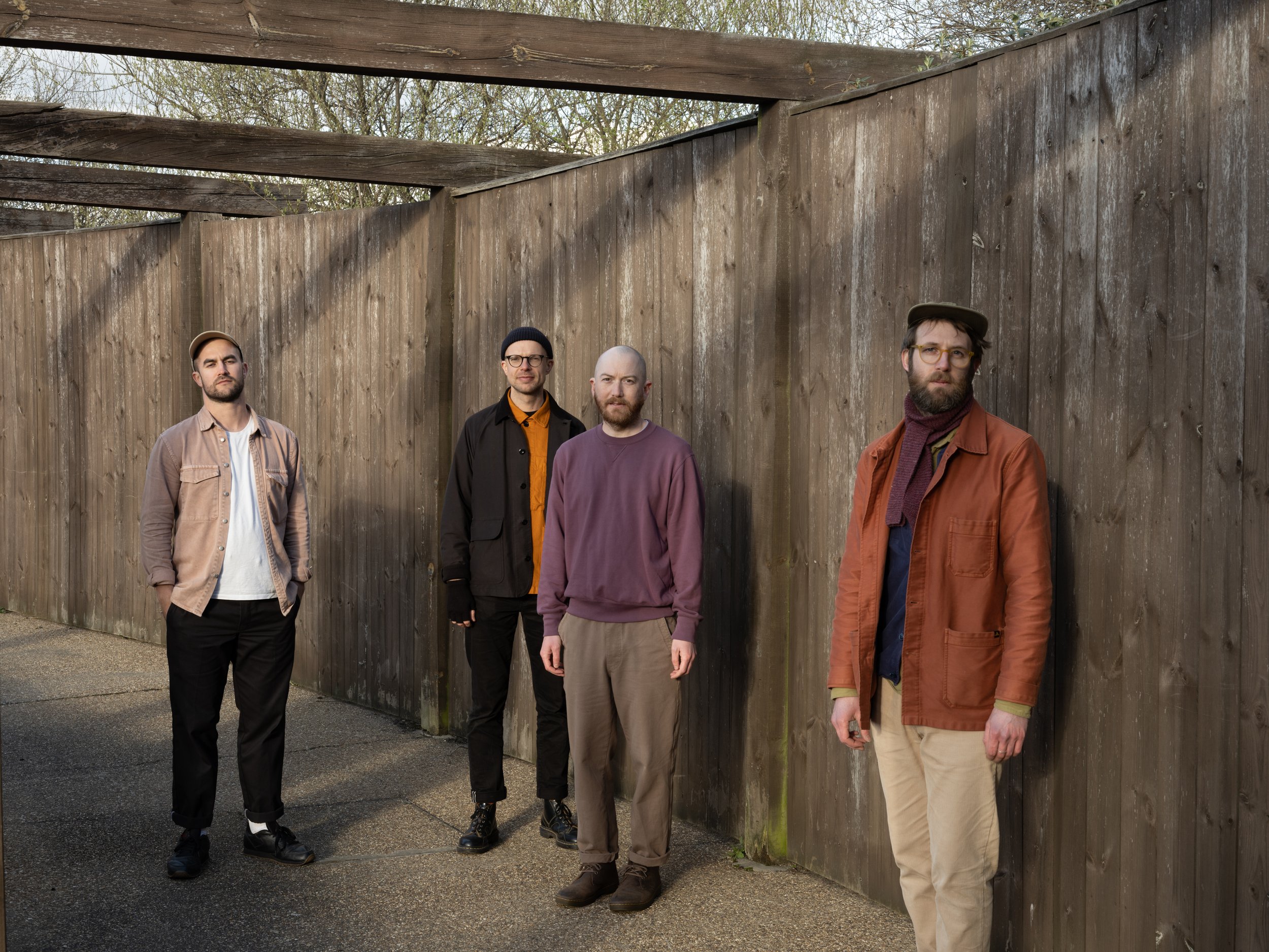 Four men standing in front of a tall wooden fence outdoors during daytime.