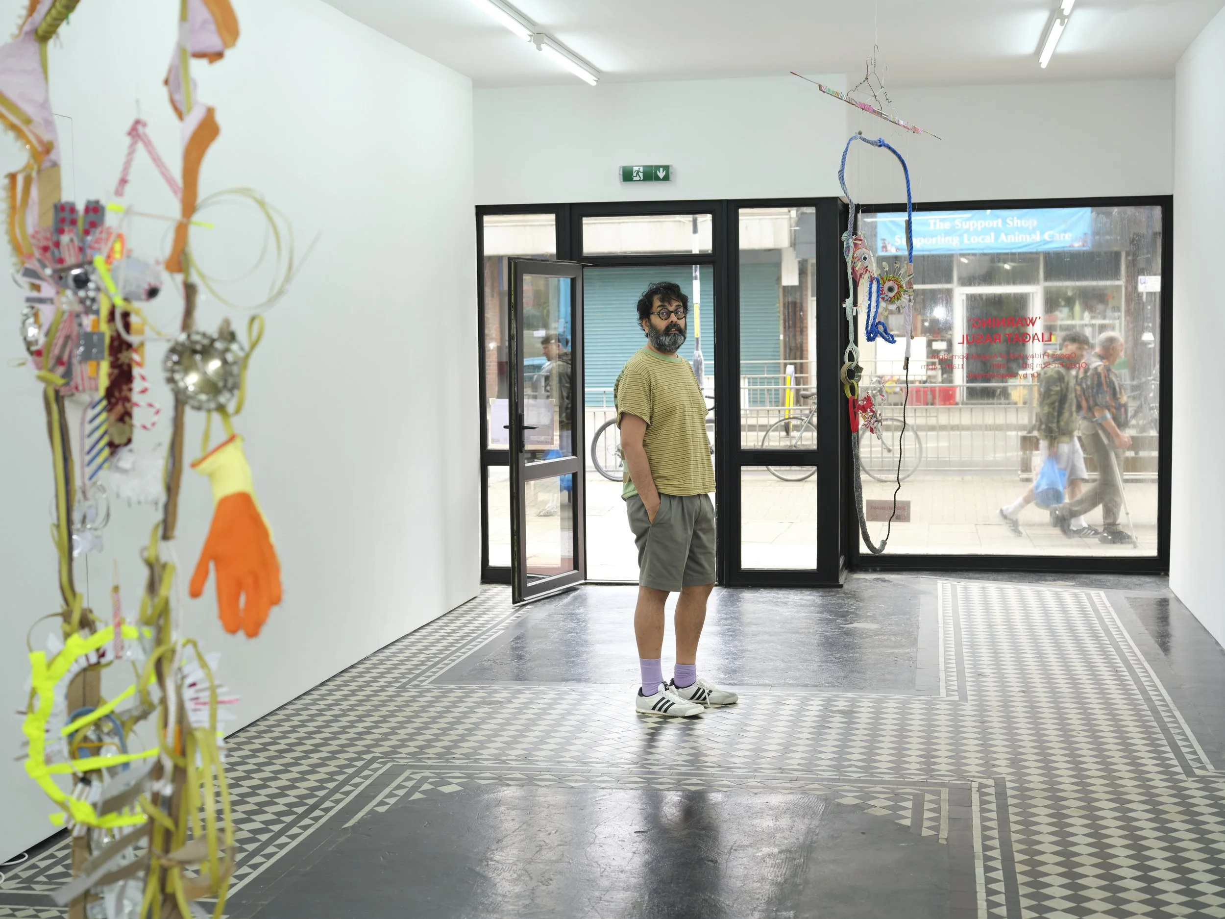 A man with glasses, a beard, wearing a yellow striped shirt, gray shorts, white sneakers, and pink socks, standing inside a store near the entrance, looking outside through the glass doors. The store has a sign in the window supporting local animal c