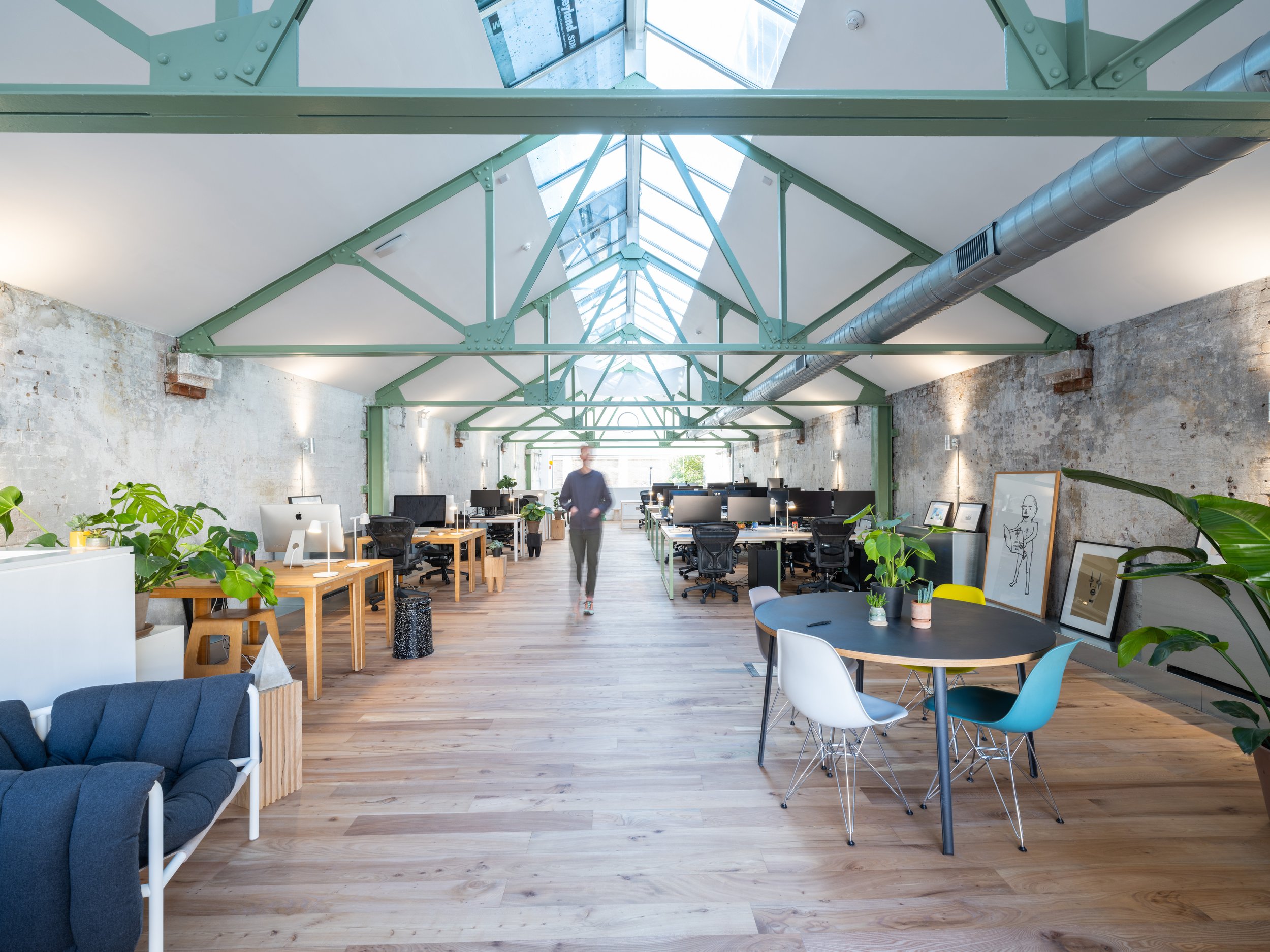 Modern office space with exposed brick walls, green metal beams, and a large skylight ceiling. There are various workstations with computers, tables, and chairs, along with some decorative plants and artwork.