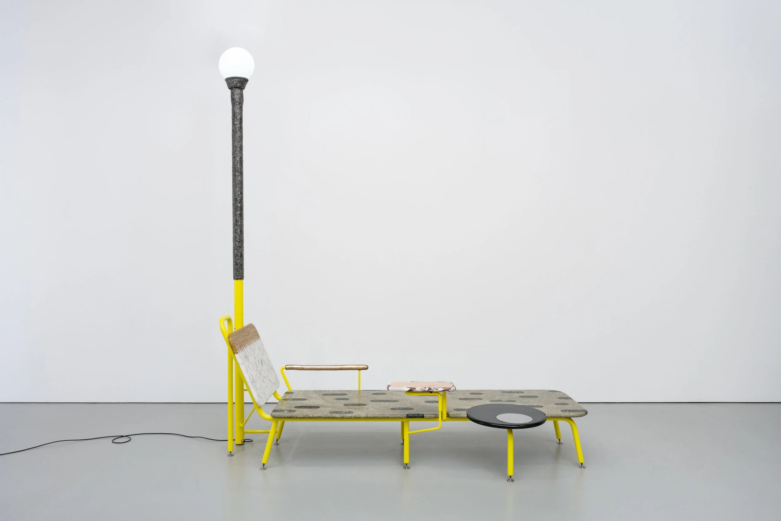 An art installation featuring a collection of furniture and objects, including a tall black metal pole with a white globe on top, a yellow chair, a small black round table, and other yellow metal structures with wooden seats, arranged against a stark