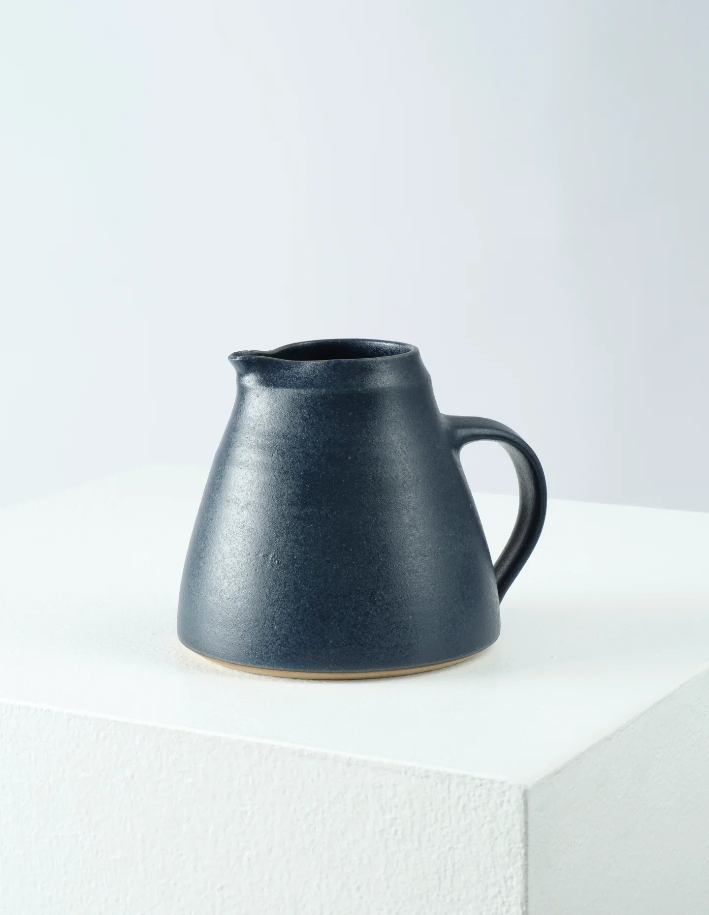 A dark blue ceramic pitcher with a matte finish placed on a white display stand against a plain light background.