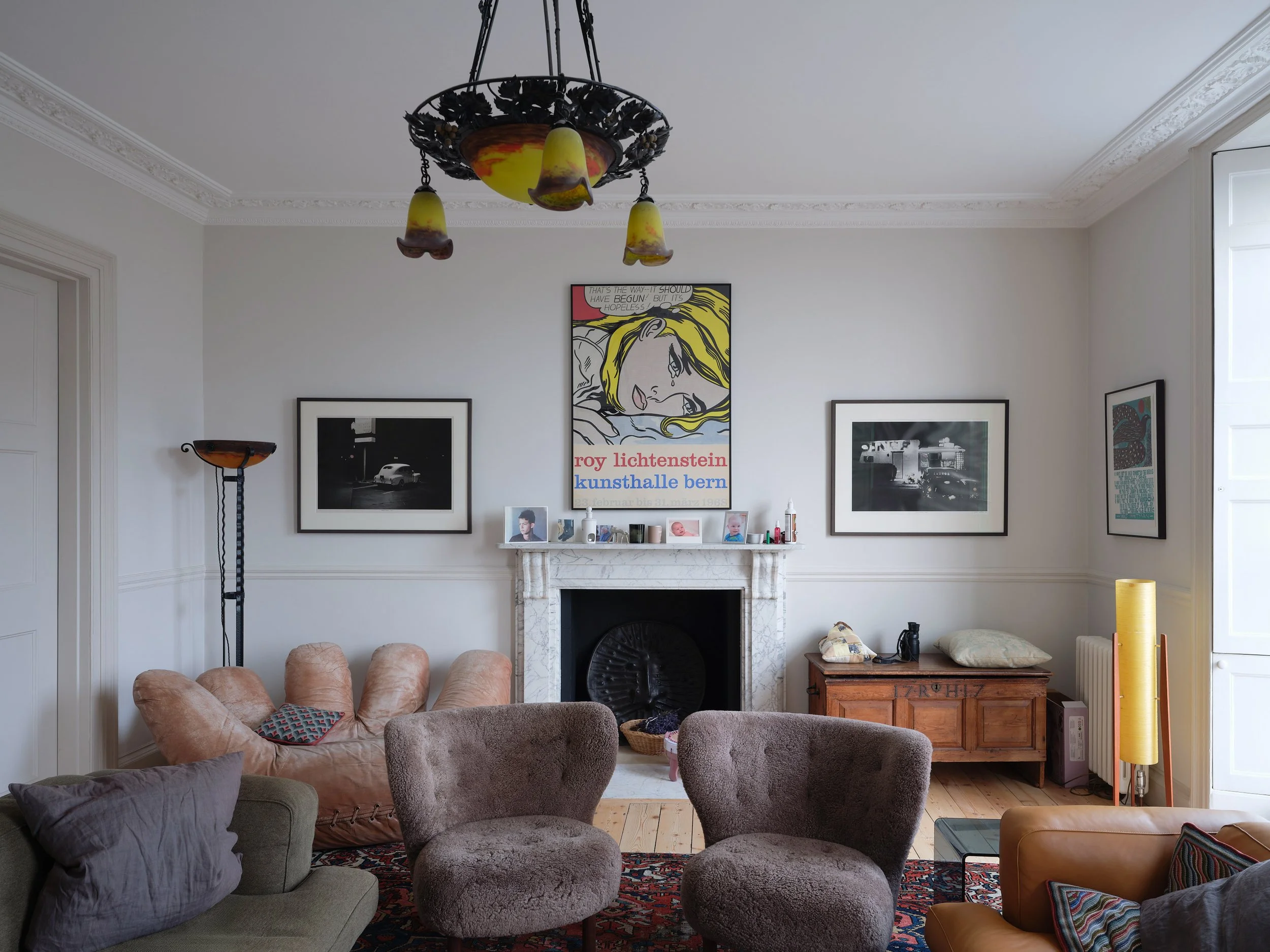 Living room with beige and gray seats, a white marble fireplace, framed artwork on the wall, a chandelier hanging from the ceiling, a window on the right, a wooden chest, and a red patterned rug.