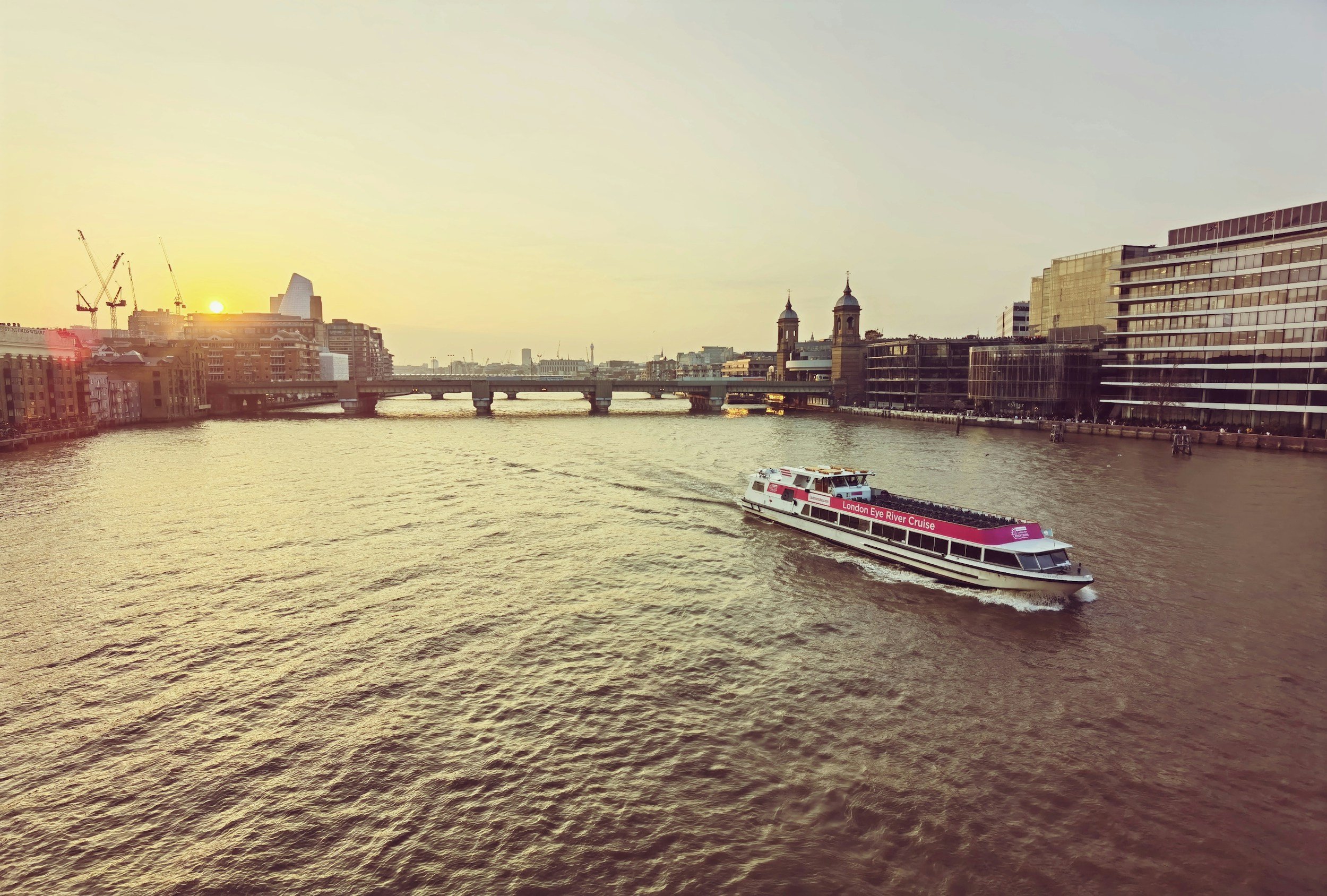 A river cruise boat on the water with a city skyline in the background during sunset.