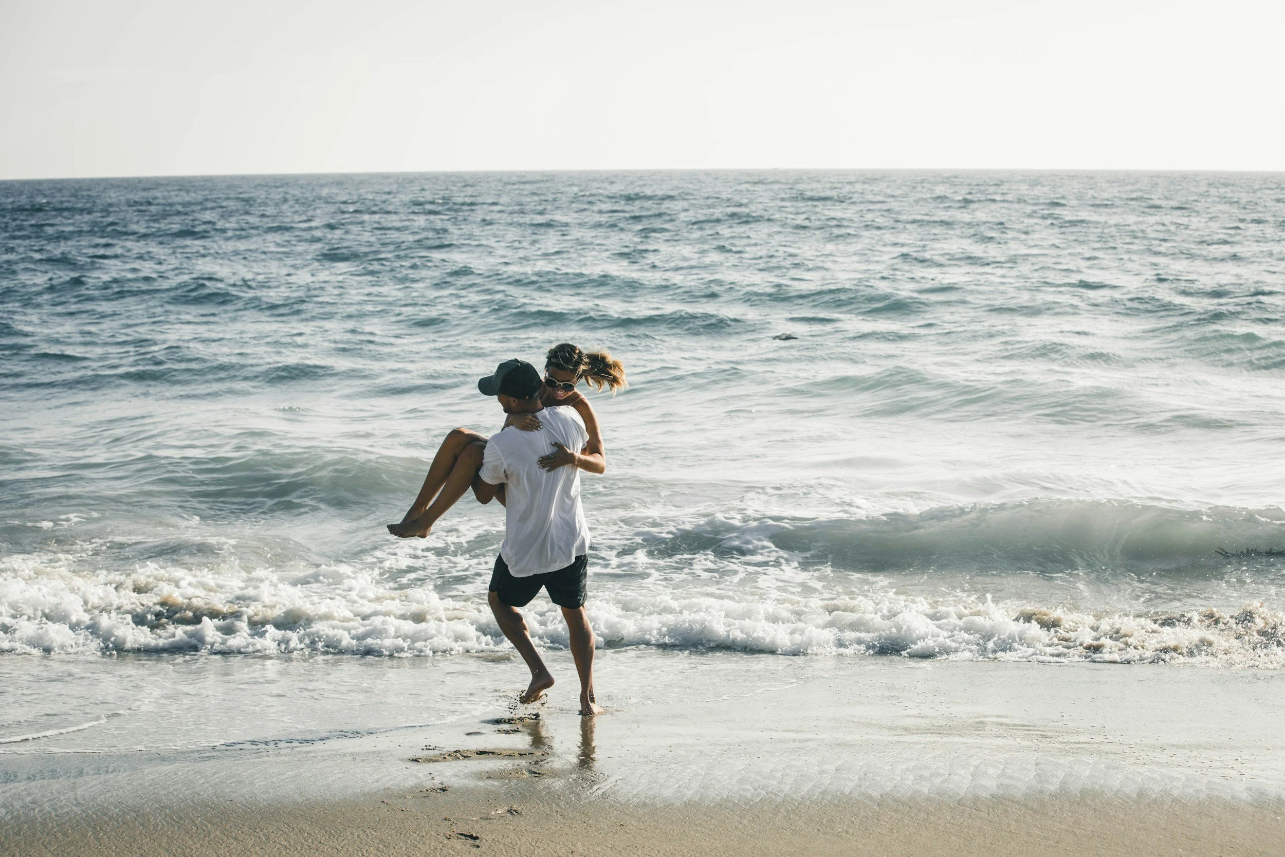 Couple at the beach, where a man is carrying a woman in his arms near the shoreline with the ocean and sky in the background.