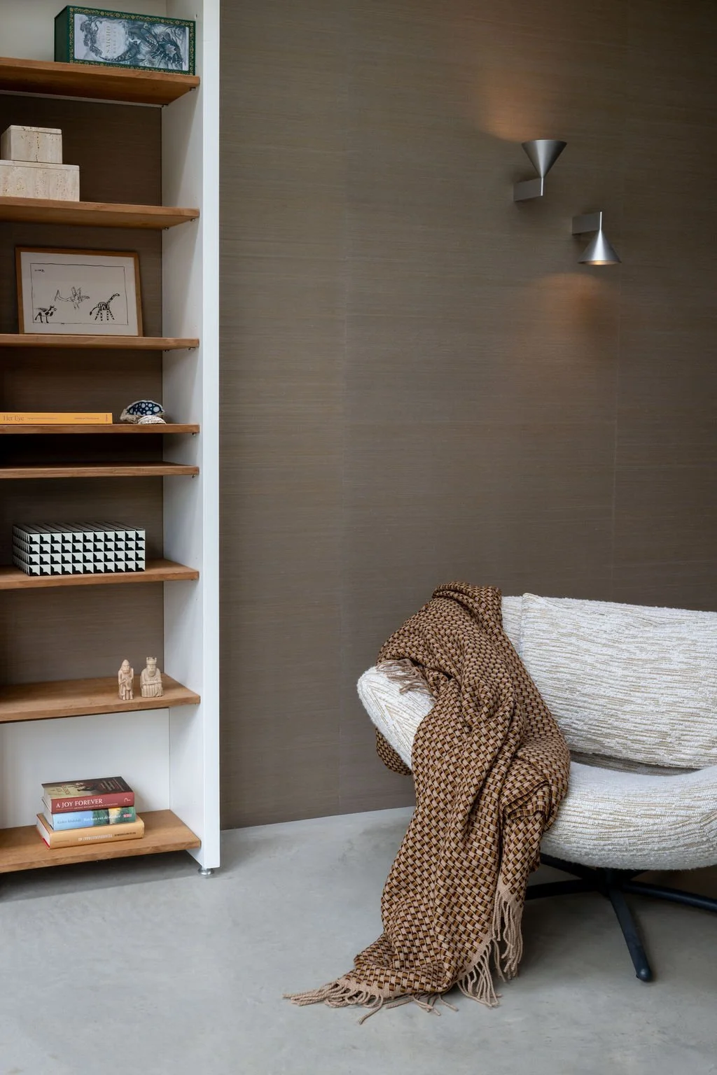 A cozy living room corner with a white textured armchair draped with a brown patterned throw, a gray wall with two modern cone-shaped wall sconces, and a white bookshelf with various decorative items and books.