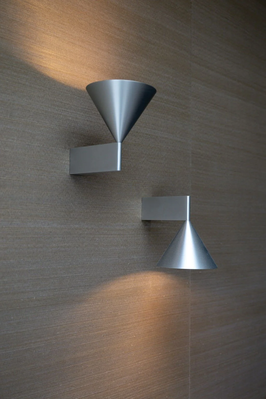 Two modern wall-mounted cone-shaped silver light fixtures on a textured beige wall, emitting soft light.