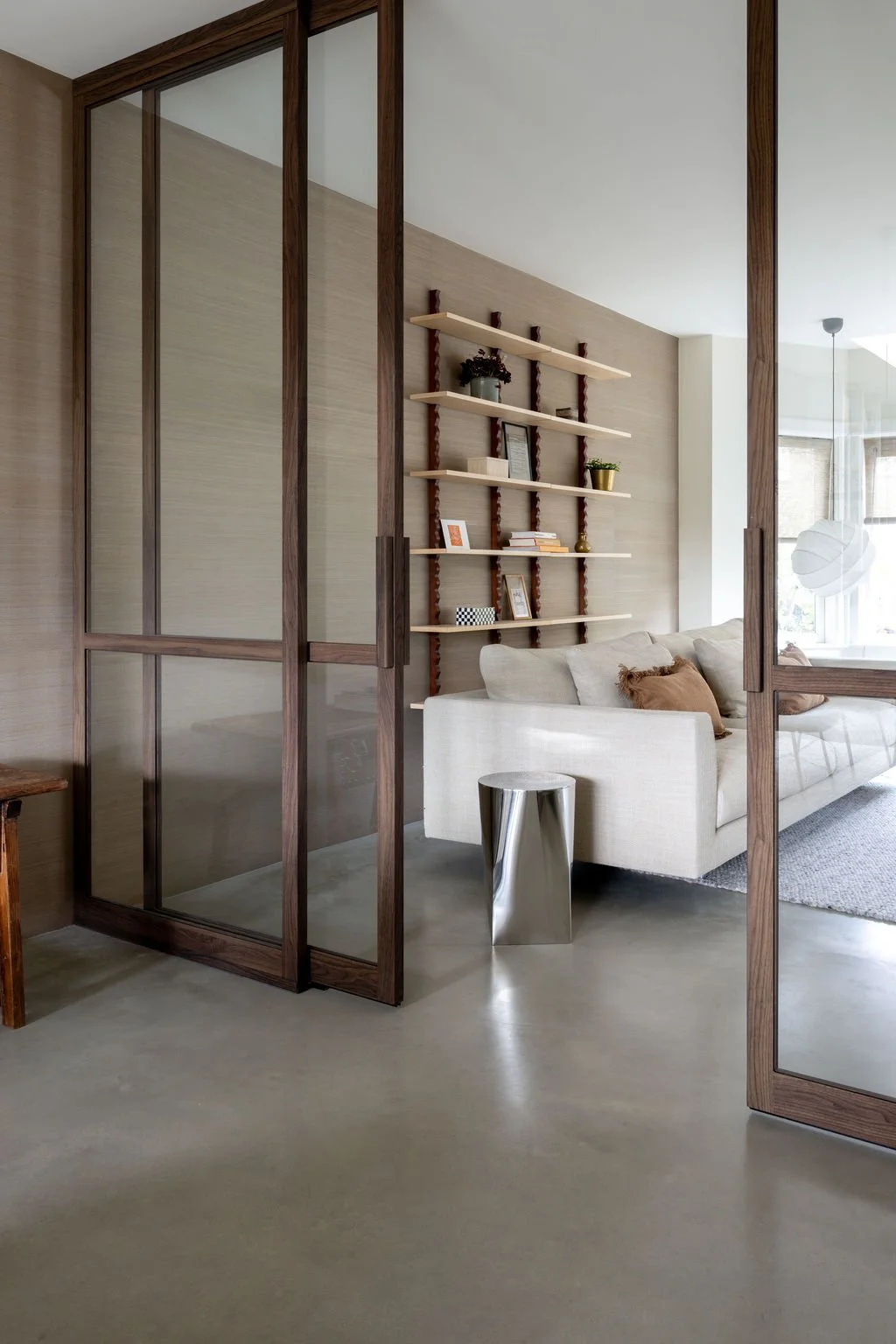 Living room interior with beige sofa, modern metallic side table, wooden and glass sliding doors, and open shelving with decorative items on textured wall.