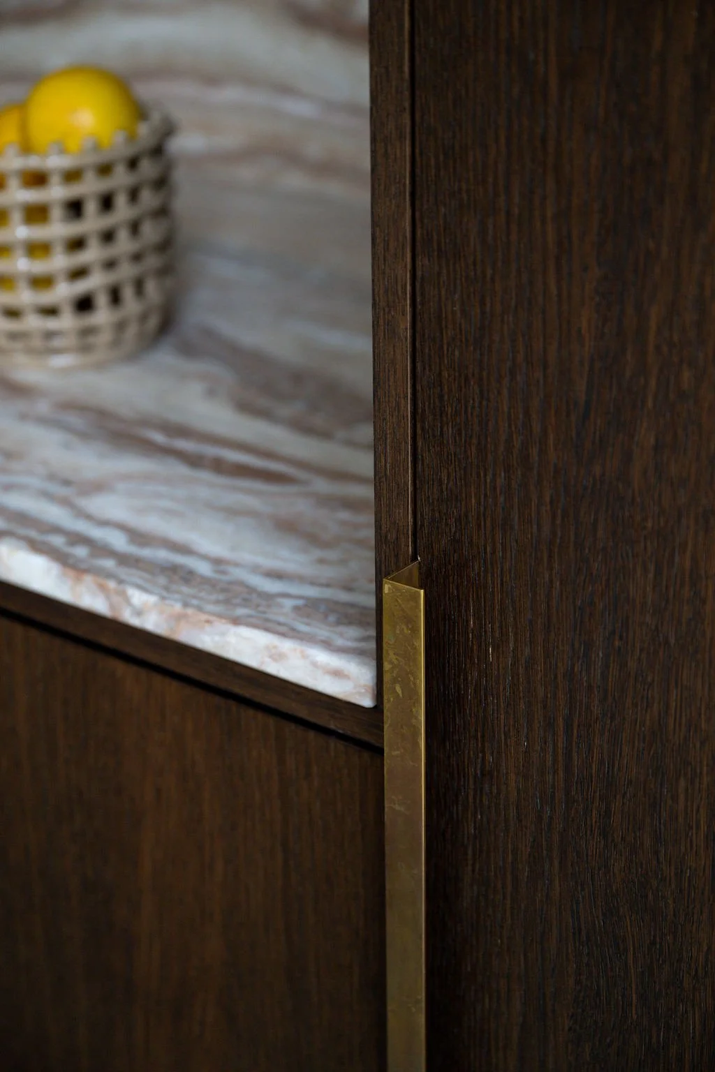 Close-up of a kitchen cabinet with a marble countertop and a brass handle, with a ceramic basket holding yellow lemons in the background.