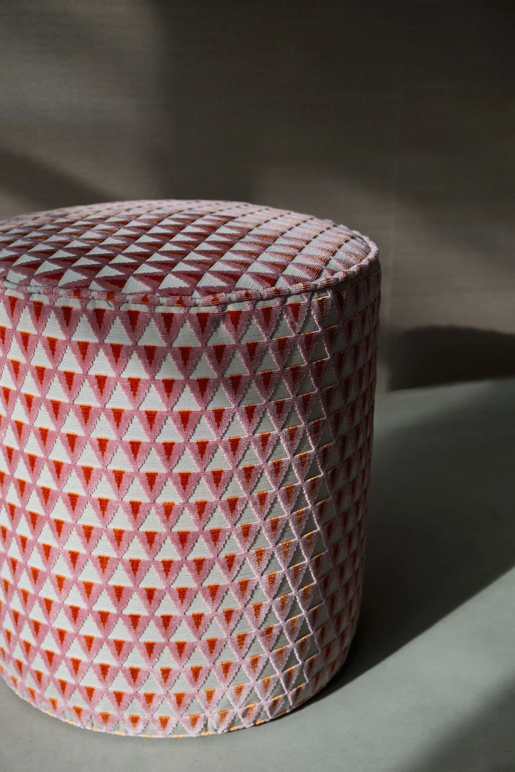 A round fabric-covered box with a geometric orange, pink, and beige pattern. It is placed on a gray surface with a shadow cast behind it.