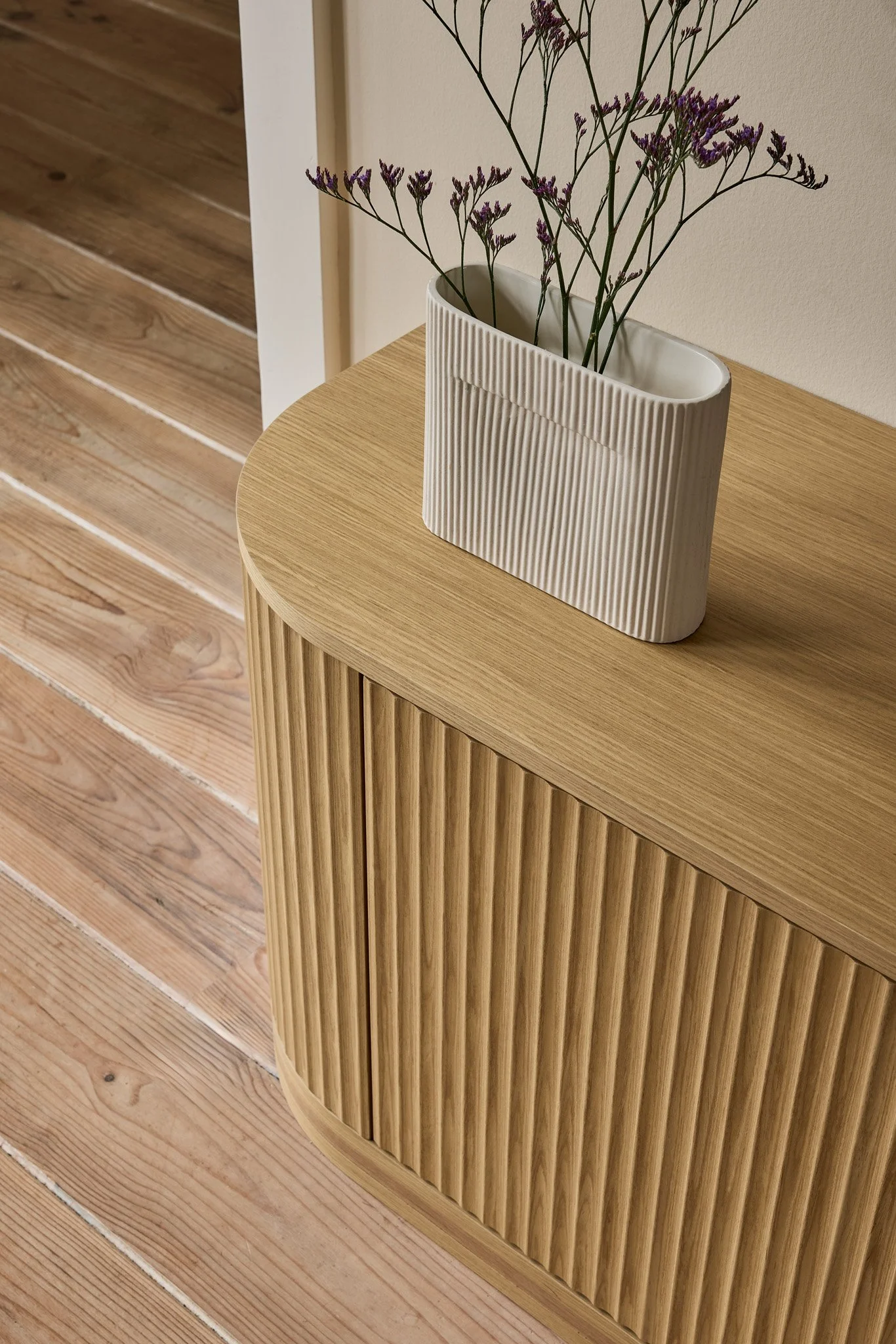 A beige ceramic vase with vertically ridged surface, containing purple flowering stems, placed on a wooden surface against a beige wall.