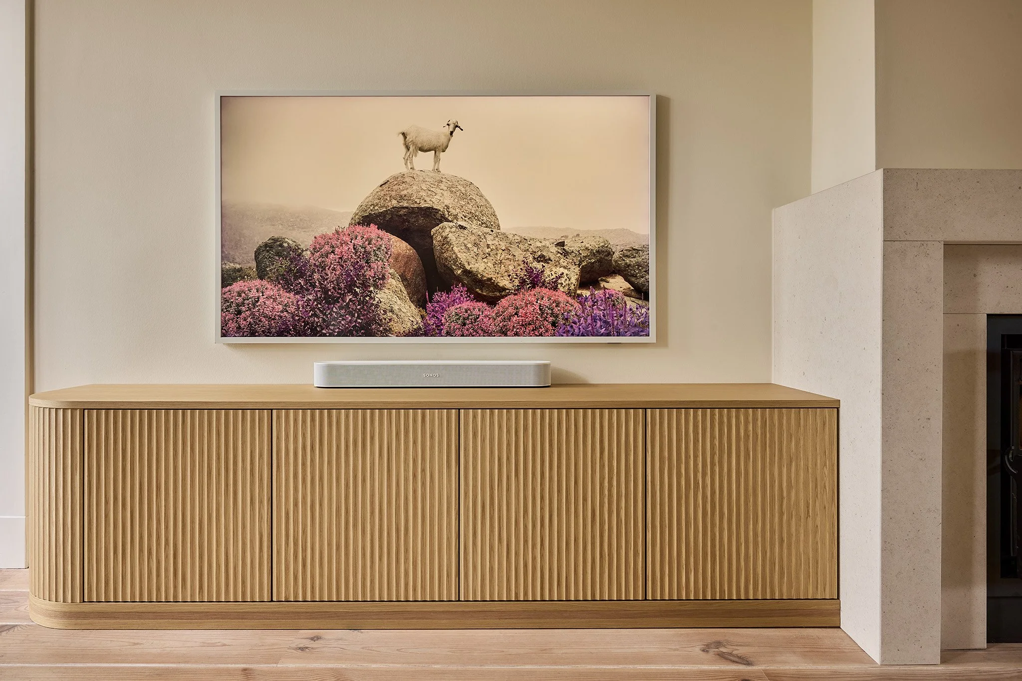 A framed picture of a goat standing on large rocks and pink and purple blooming plants, mounted above a wooden cabinet with vertical slats in a living room.
