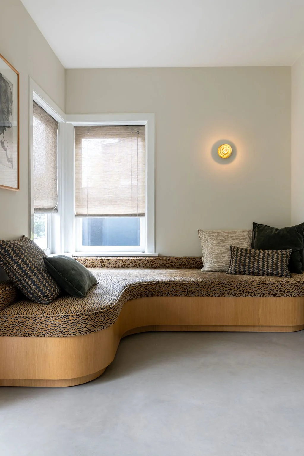 A cozy corner of a room with a built-in cushioned bench under a window, decorated with black and beige pillows. The window has light-colored blinds, and there is a small yellow wall sconce on the cream-colored wall. Part of a framed artwork is visibl