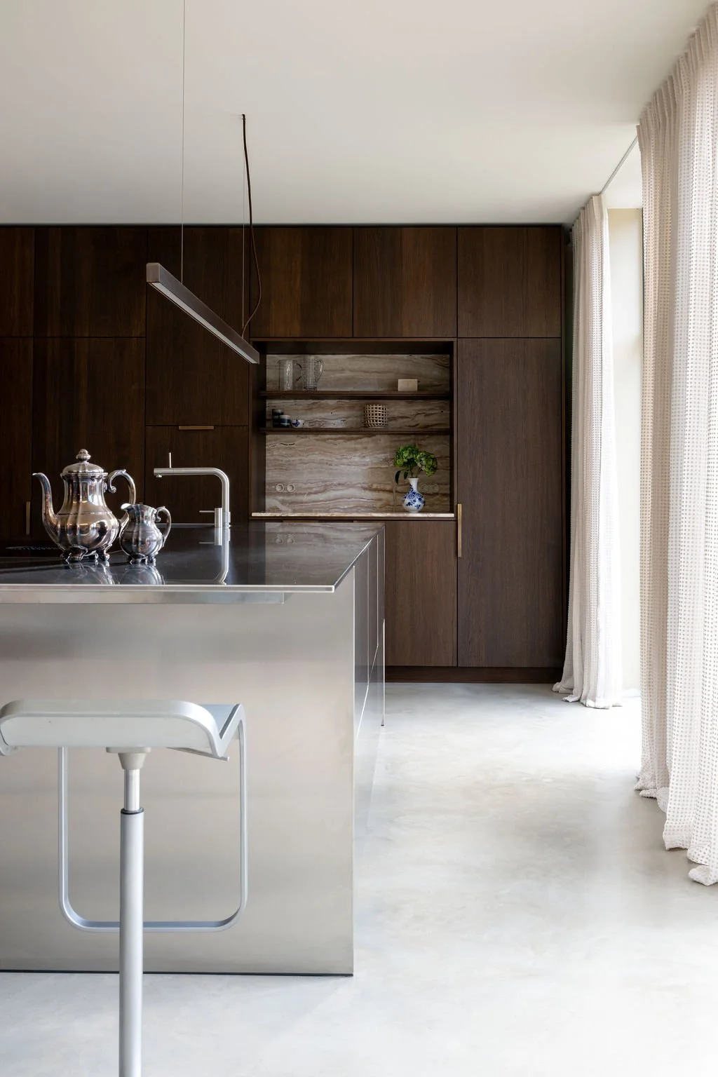 RVS keukeneiland met donker hout en messing accenten, gordijnen met structuur die spelen met het licht.