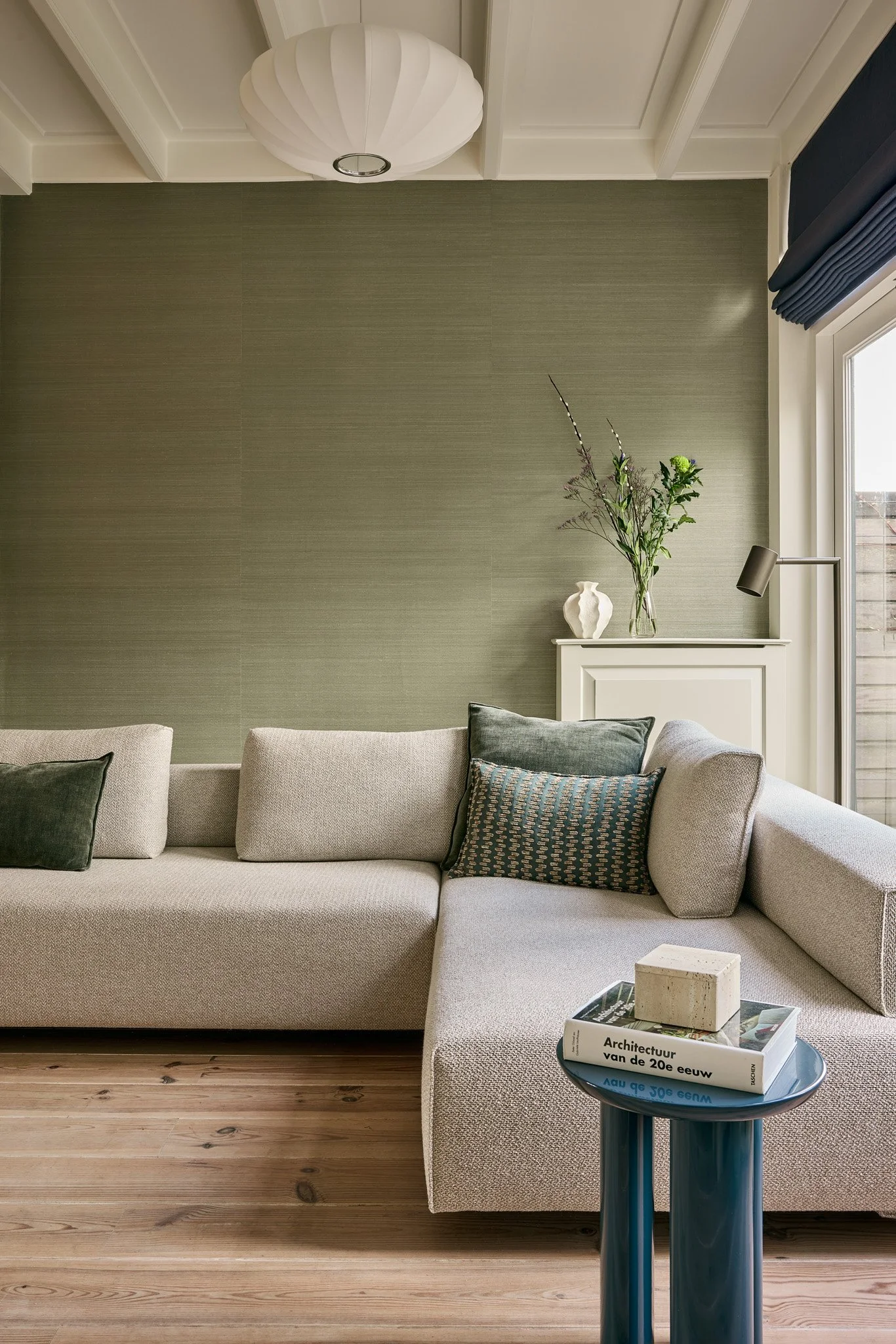 Living room with a beige sectional sofa with dark and patterned throw pillows, a small round side table with stacked books, a modern ajoutature, and a beige wall with a decorative shelf holding a white vase and a glass vase with flowers. A window wit