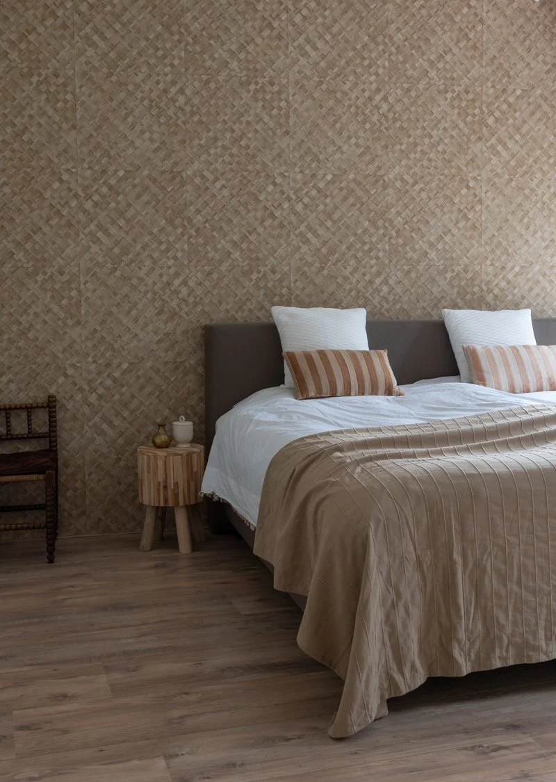 A neatly made bed with white and striped pillows, a beige bedspread, and a woven bamboo wall behind it in a bedroom with wooden flooring.