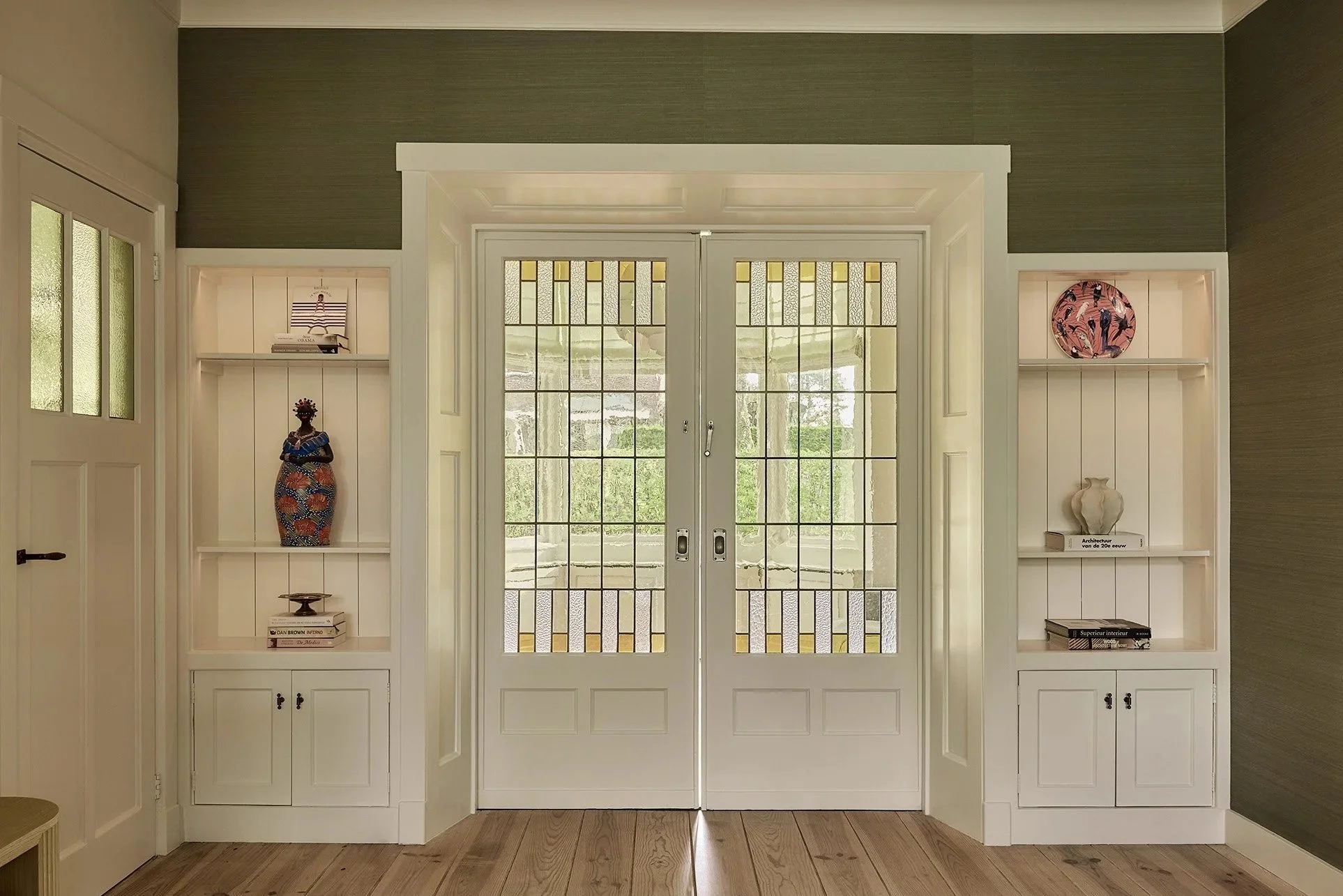 Living room with open glass double doors, flanked by built-in white shelves with decorative items and books, on a wood floor and green textured wall.