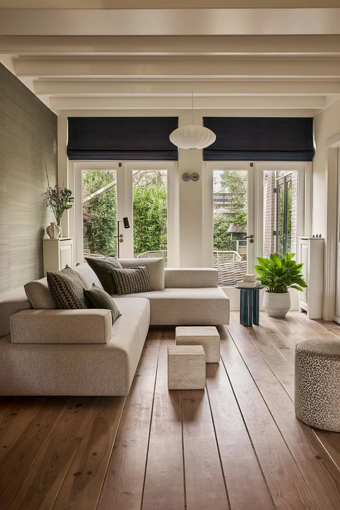 Living room with beige sofa, throw pillows, a white side table, potted plant, and wooden flooring, with large windows and a view of greenery outside.