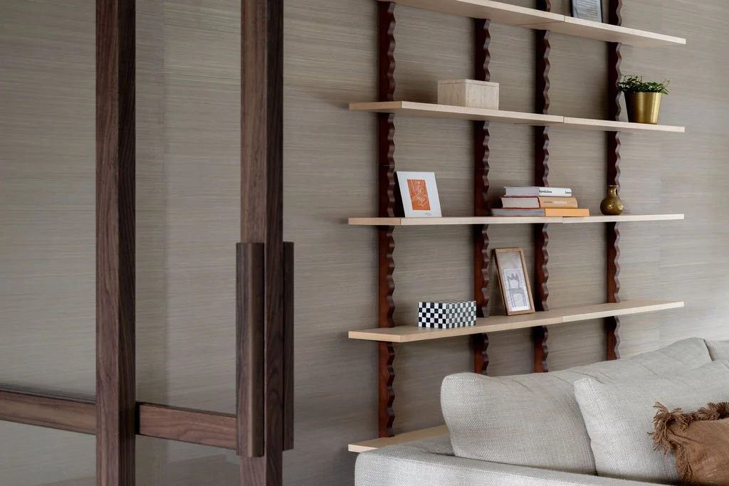 A modern living room with open wooden shelves on the wall, decorated with books, picture frames, a small vase, and a potted plant. Part of a beige sofa is visible in the bottom right corner.