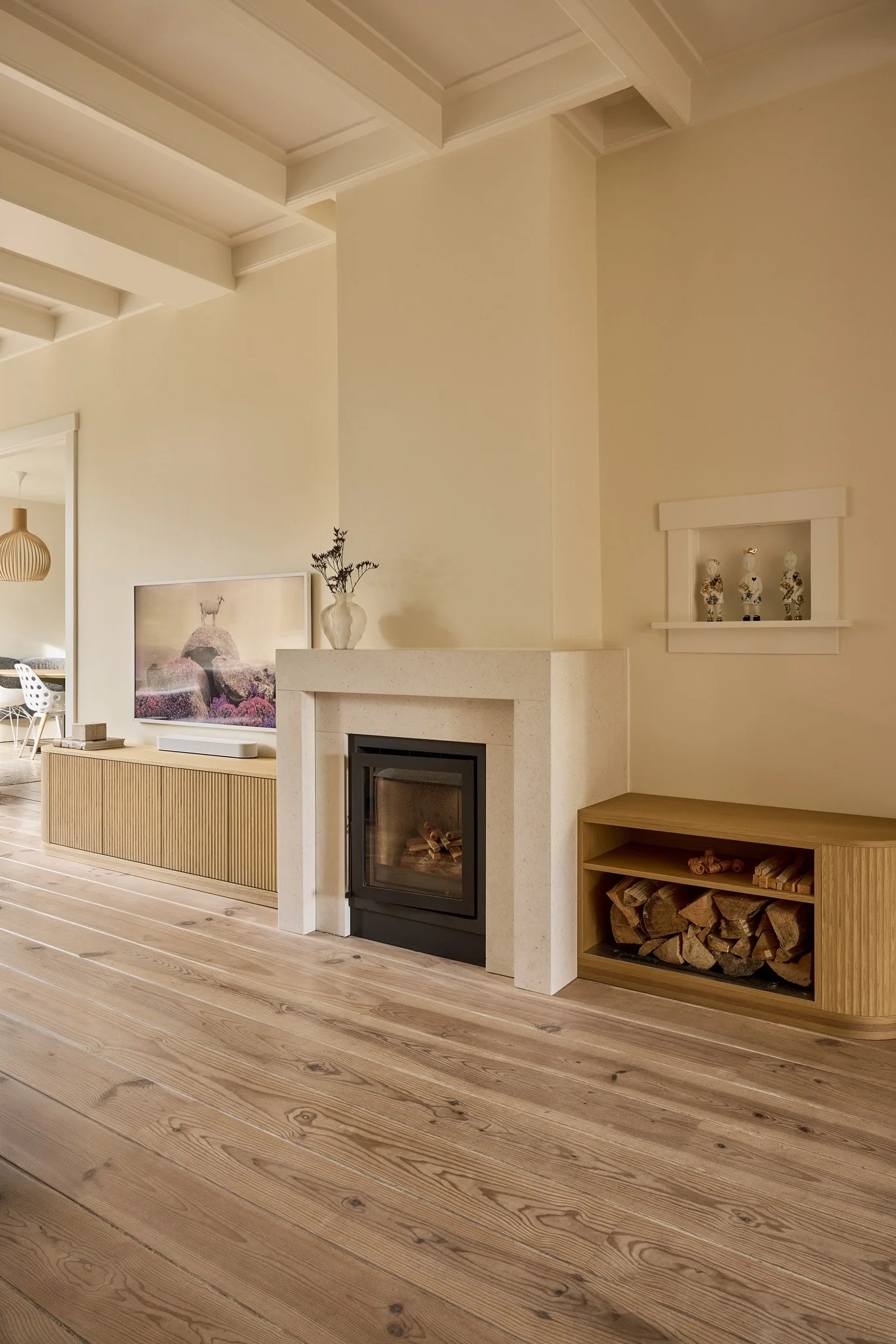 Living room with wood flooring, a fireplace with a white mantle, a wooden cabinetry with logs stored inside, a framed landscape picture, a vase with greenery, and a built-in shelf with decorative items, in a neutral-colored space.