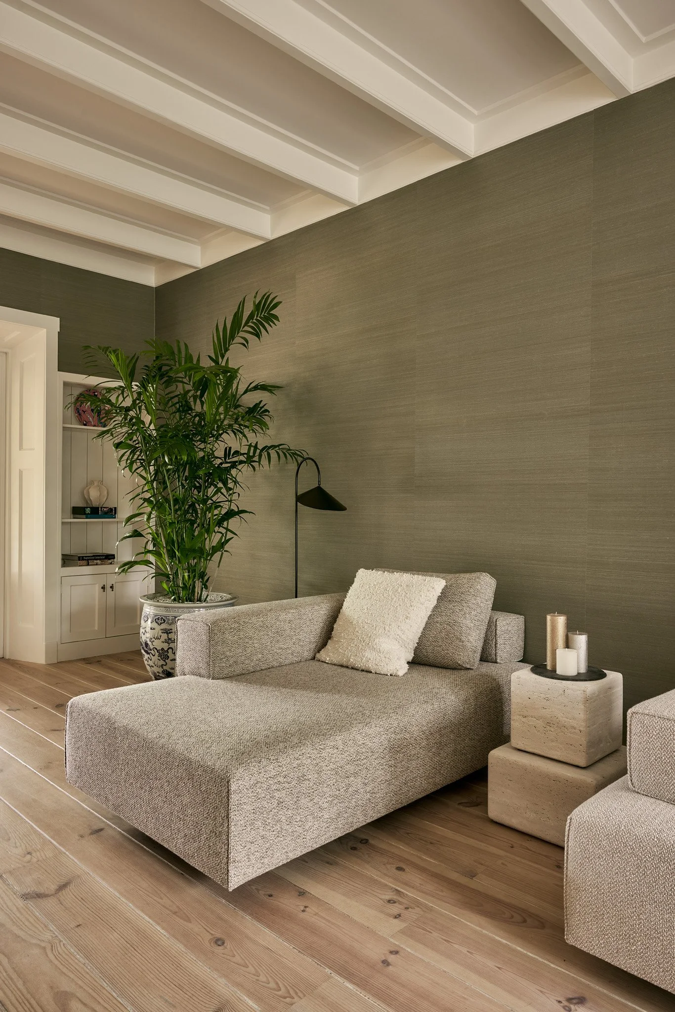 Living room with a beige sectional sofa, white textured pillows, a black floor lamp, a small round table with candles, a large potted plant, built-in white shelves, and brown wooden flooring, against a taupe textured wall with a coffered ceiling.