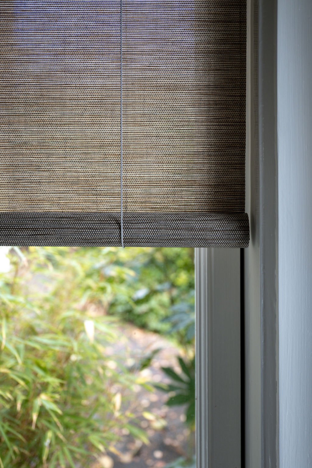 A close-up of a window with a gray pull-down shade partially lowered, showing greenery outside, including plants and bushes.