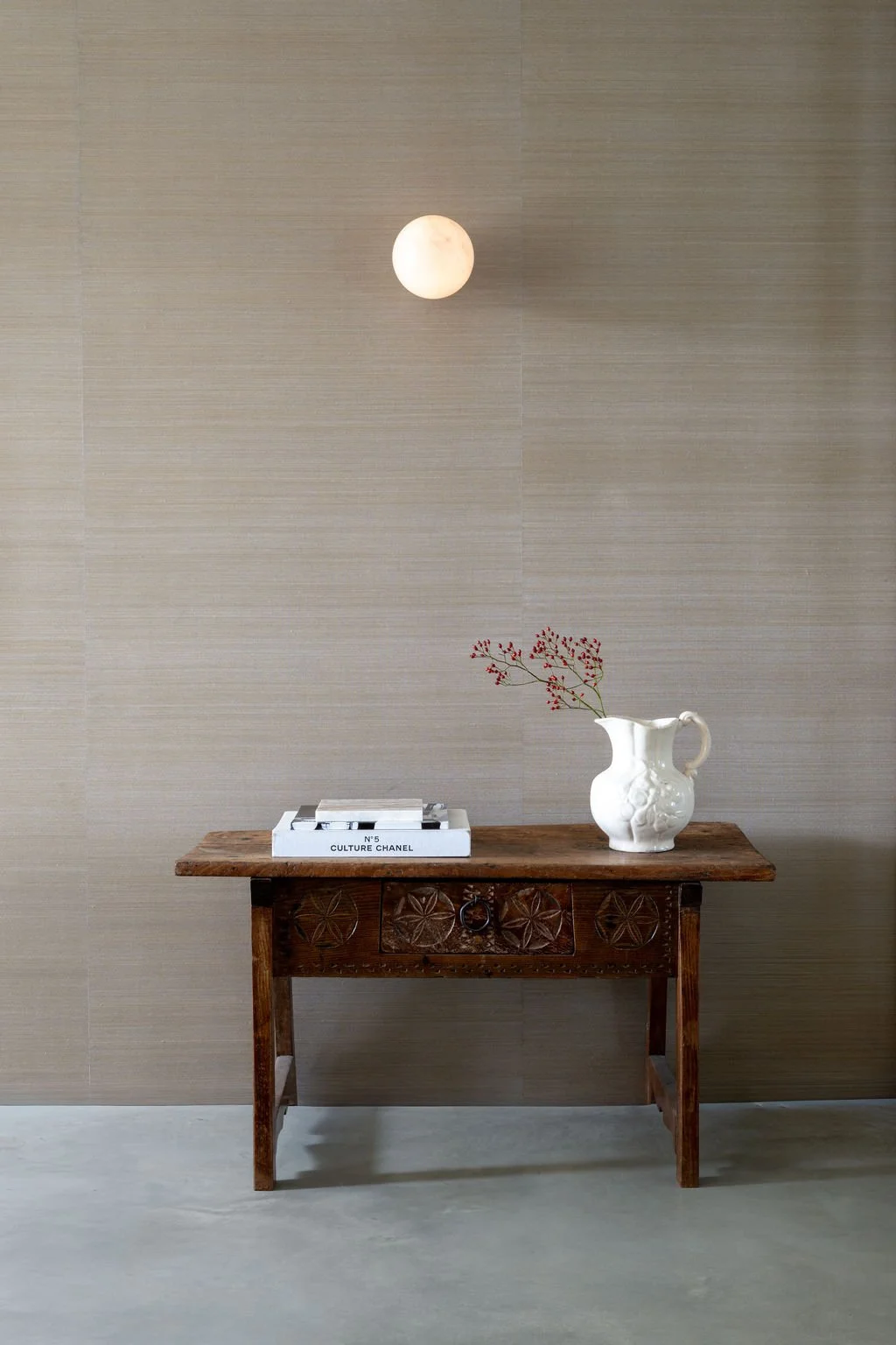 A small wooden table with carved details holds a white ceramic vase with red berries and a white box with black text, against a beige textured wall with a round ceiling light above.