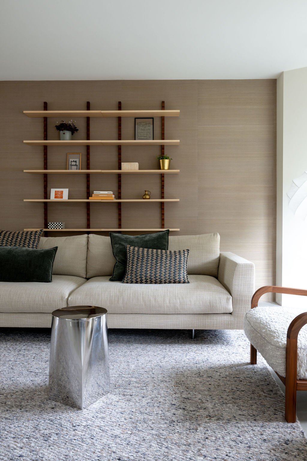 Modern living room with beige sofa, patterned pillows, metallic side table, wall-mounted wooden shelves with decorative items, and a textured rug.