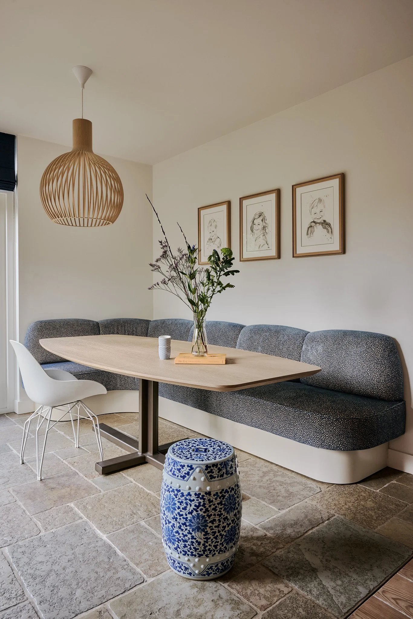 A dining area with a wooden table, a mixed material banquette seating with a dark fabric cushion, and three framed children's sketches on the wall. There is a flower arrangement on the table, a white and blue ceramic stool on the stone tile floor, an