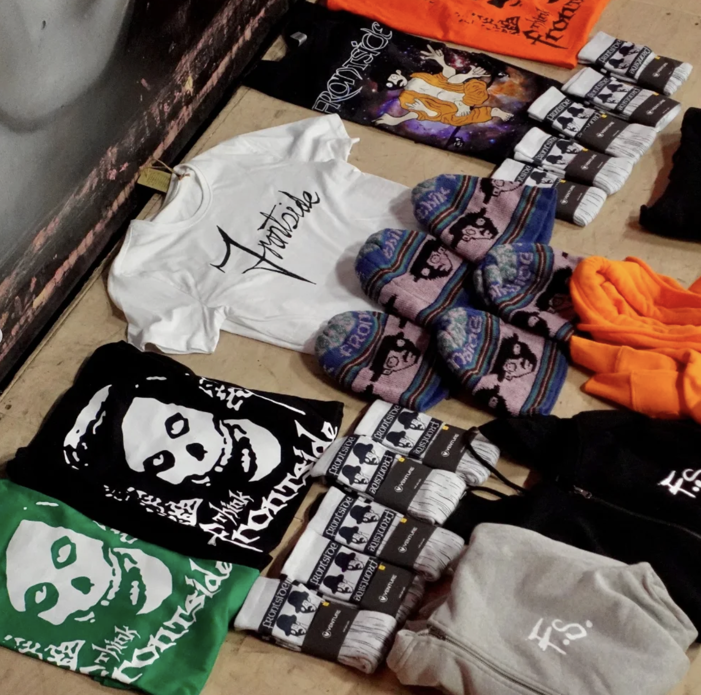 Clothing and accessories featuring the clothing brand 'Pleasures' displayed on a table, including T-shirts, socks, beanies, and a tote bag, with some items showing a skull design.