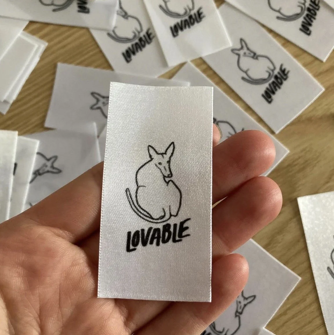 A hand holding a fabric label with a drawing of a sitting fox above the word "LOVABLE". Several similar labels are spread out on a wooden surface.