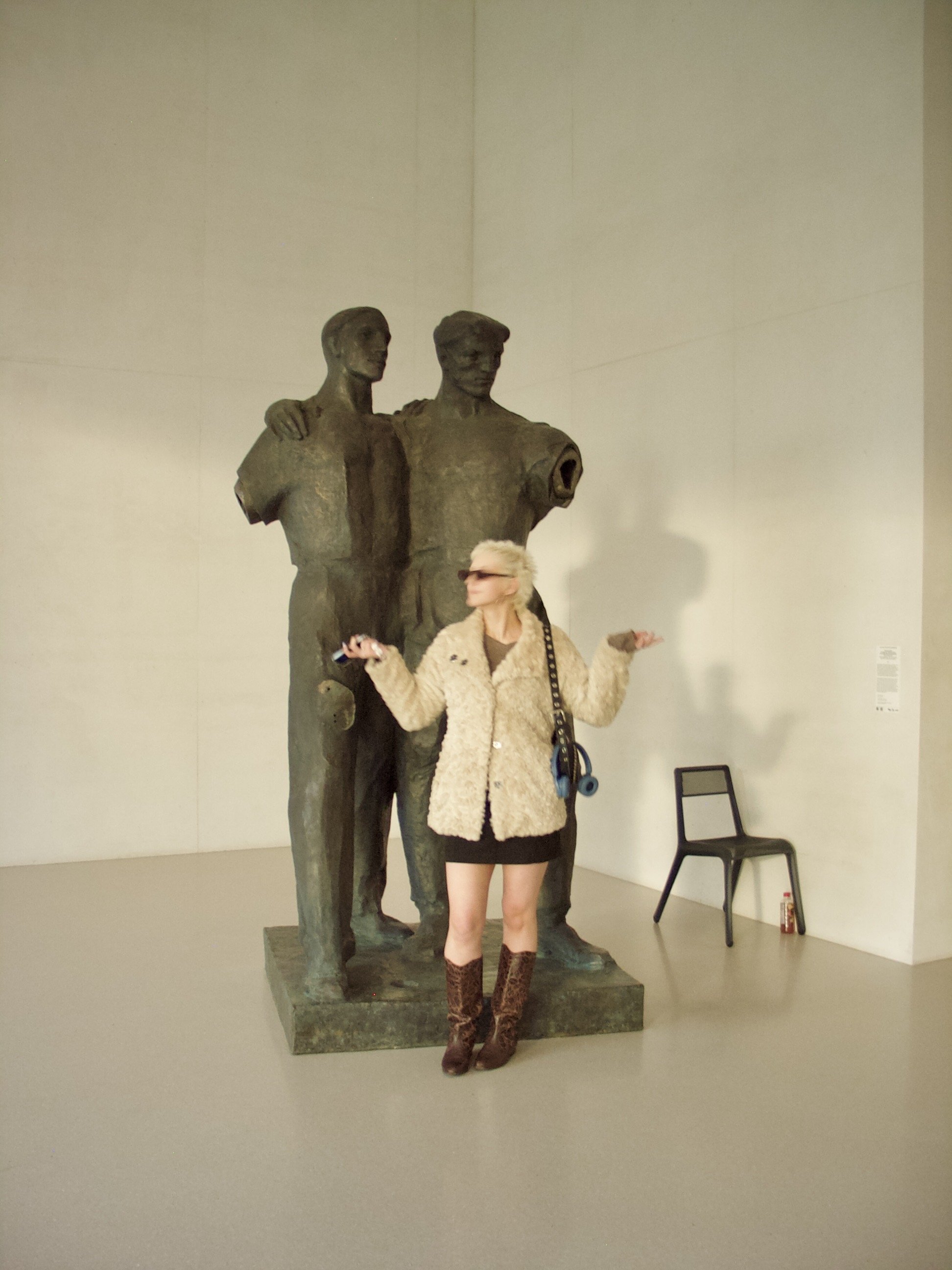 A woman with blonde hair, wearing sunglasses, a beige fur coat, a black skirt, and leopard print boots, is standing in front of a bronze sculpture of two military figures, with her arms raised in a gesture of shrugging in an art gallery.