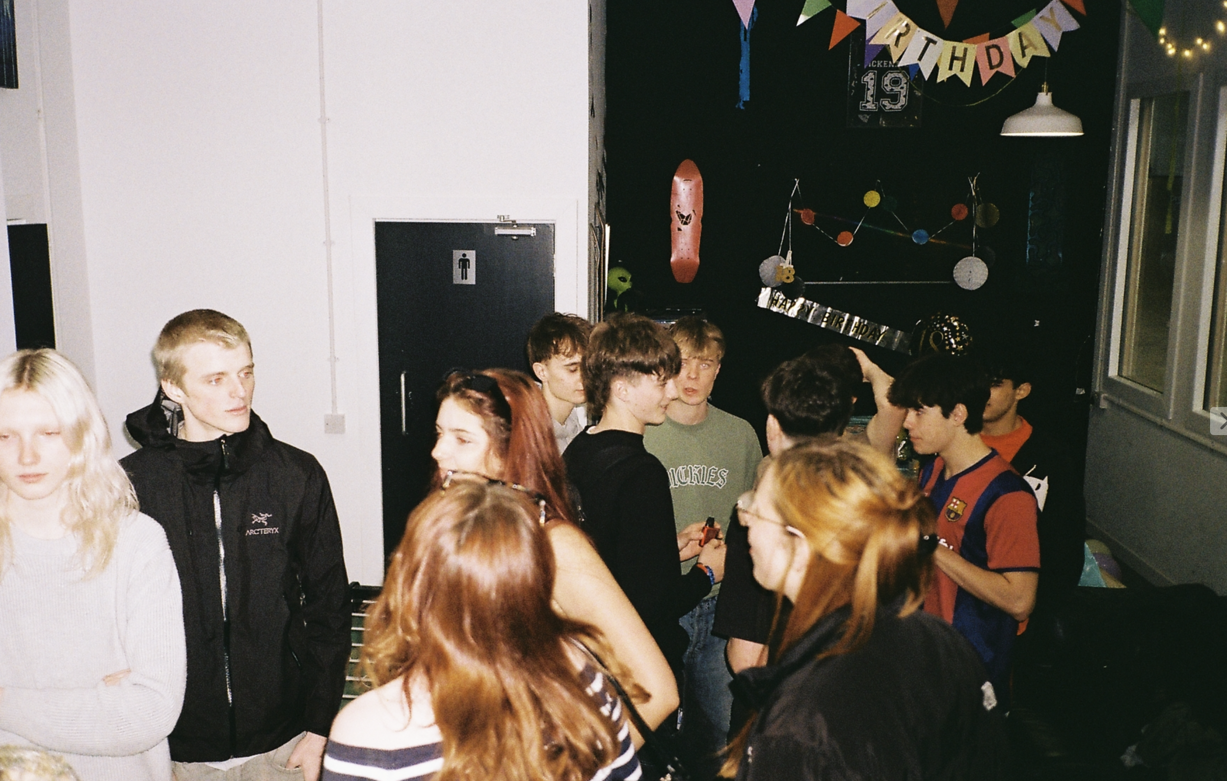 Group of young people at a birthday party with colorful decorations, including a 'Happy Birthday' banner and balloons.