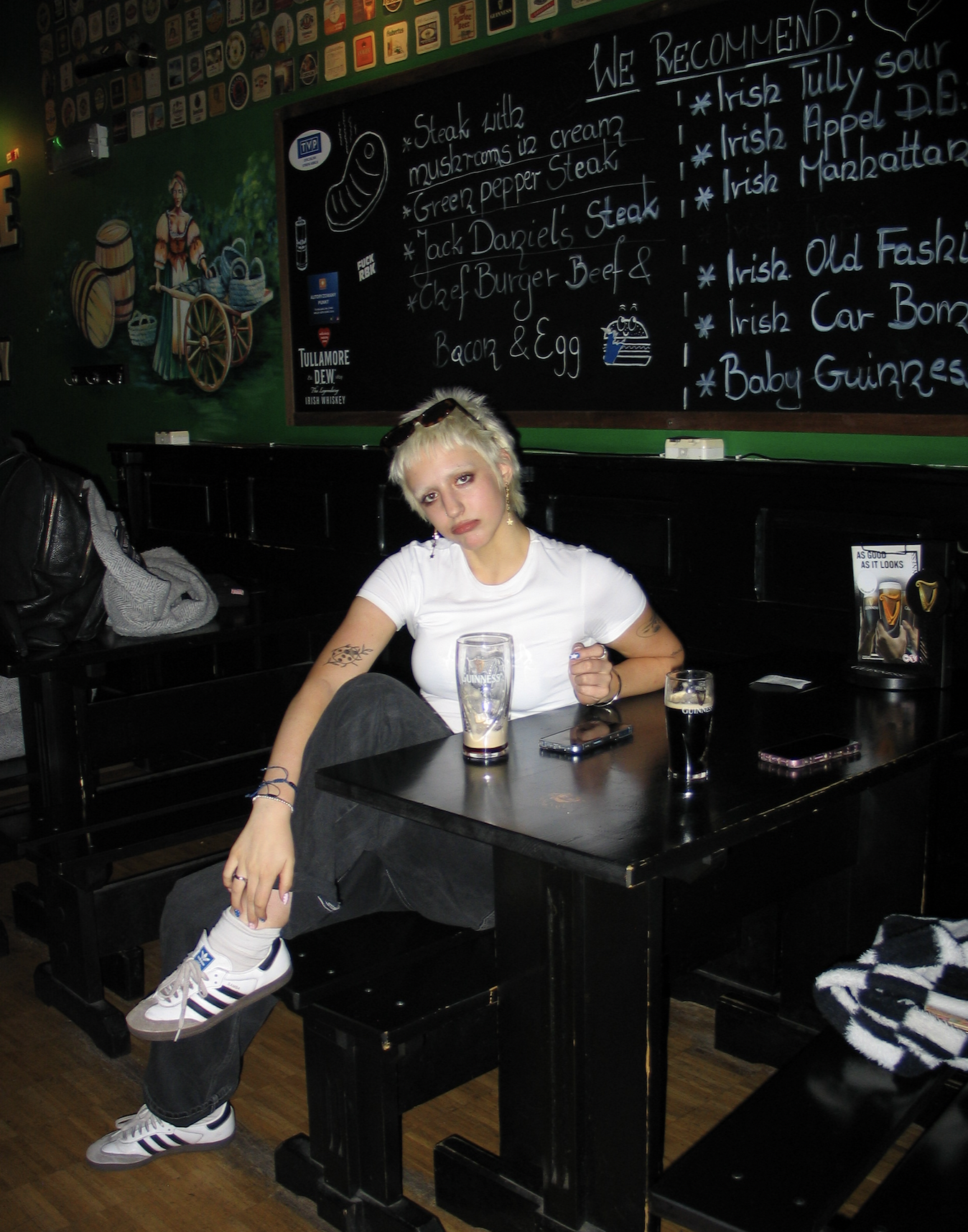 A woman with short platinum blonde hair, wearing a white t-shirt, black pants, and white sneakers with black and purple stripes, sitting at a black pub table inside a bar or restaurant. She has tattoos on her arms and is looking at the camera with a neutral expression. On the table are a tall Guinness glass, a shorter glass, a smartphone, and some personal items. The background features a chalkboard menu with Irish dishes, a painted mural of a woman in traditional Irish attire pushing a cart, and various beer coasters and advertisements on the walls.