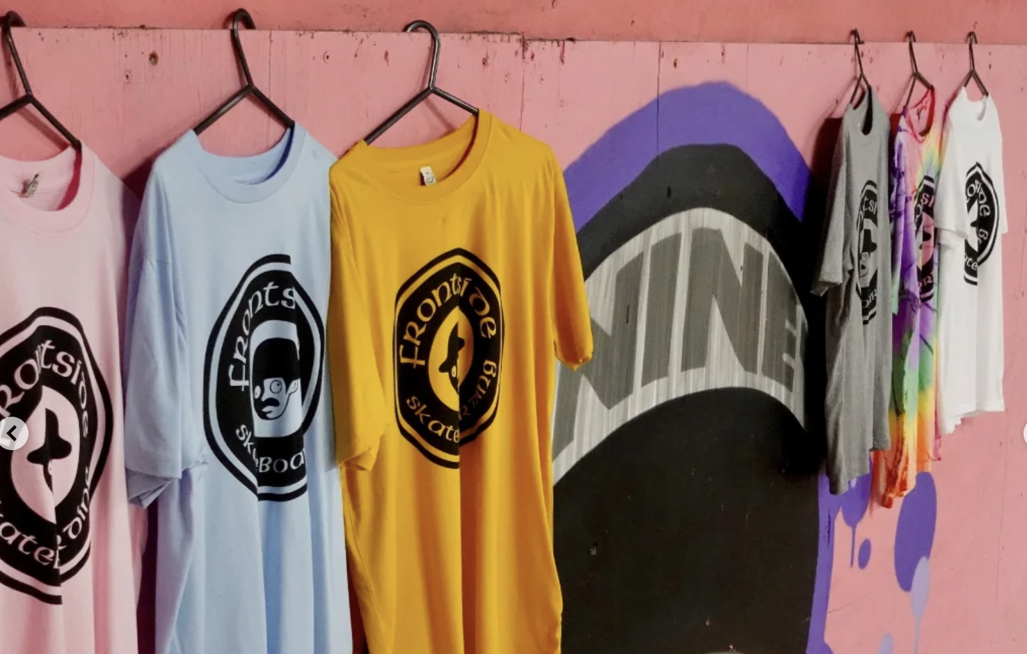 A row of colorful T-shirts hanging on a pink wall with purple and black graffiti art in the background.