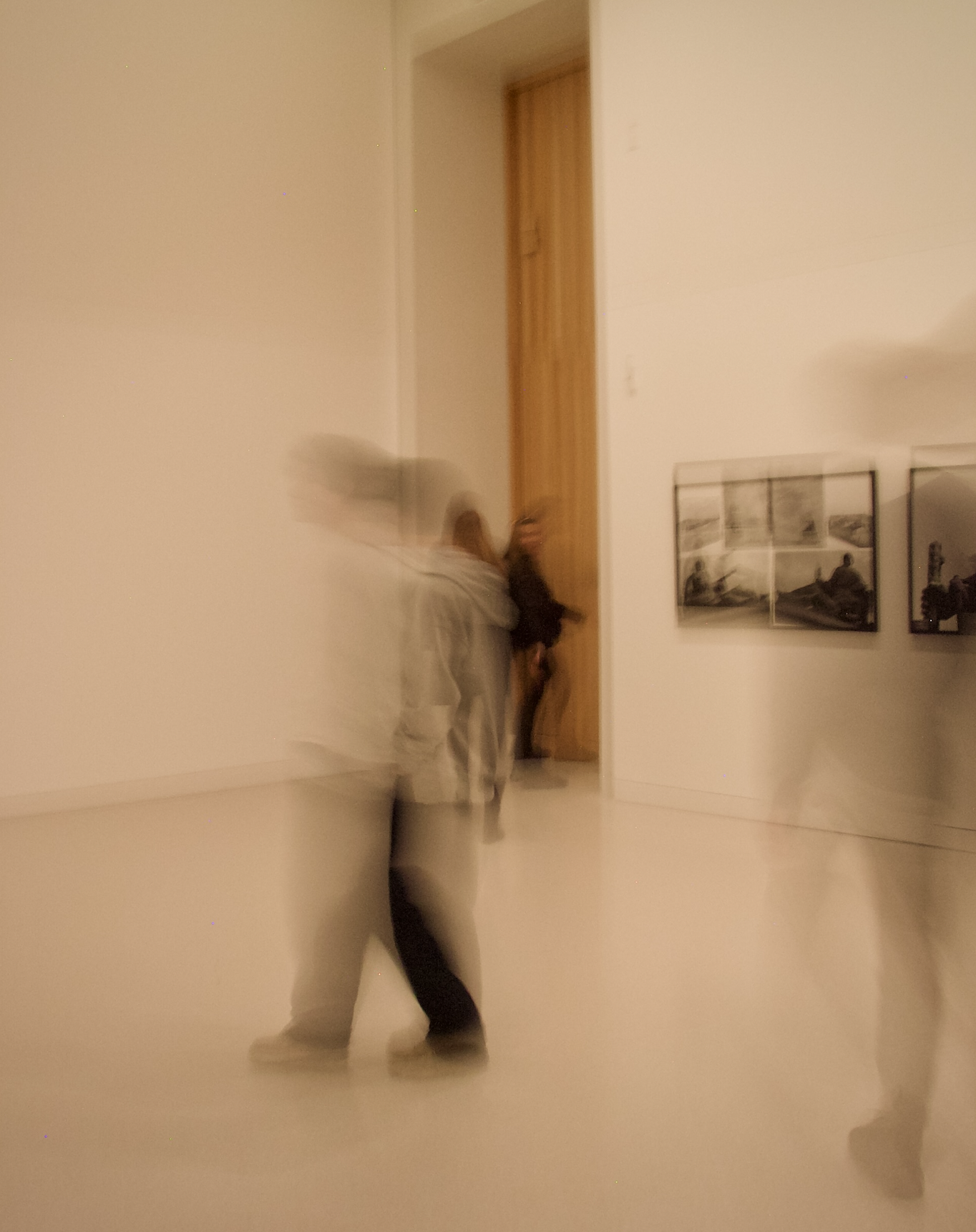 A person dressed in light-colored clothing and black pants is leaning against a wall in an art gallery or museum, with framed black-and-white photographs hanging on the wall. The photo appears to have motion blur.