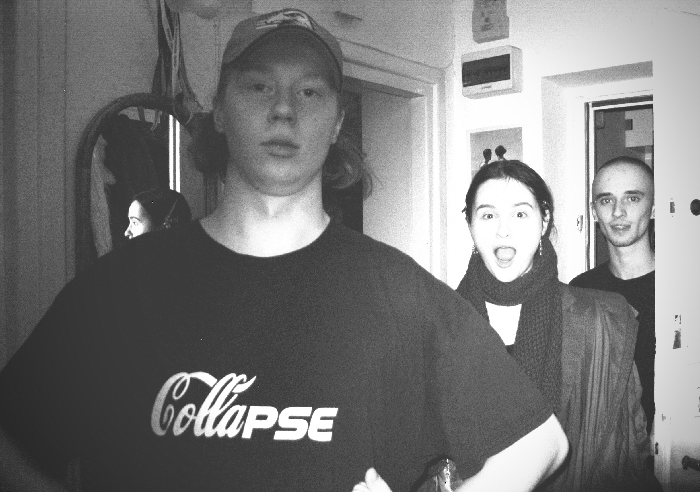 Black and white photo of three young people standing inside a room, with a mirror and wall decorations visible in the background.