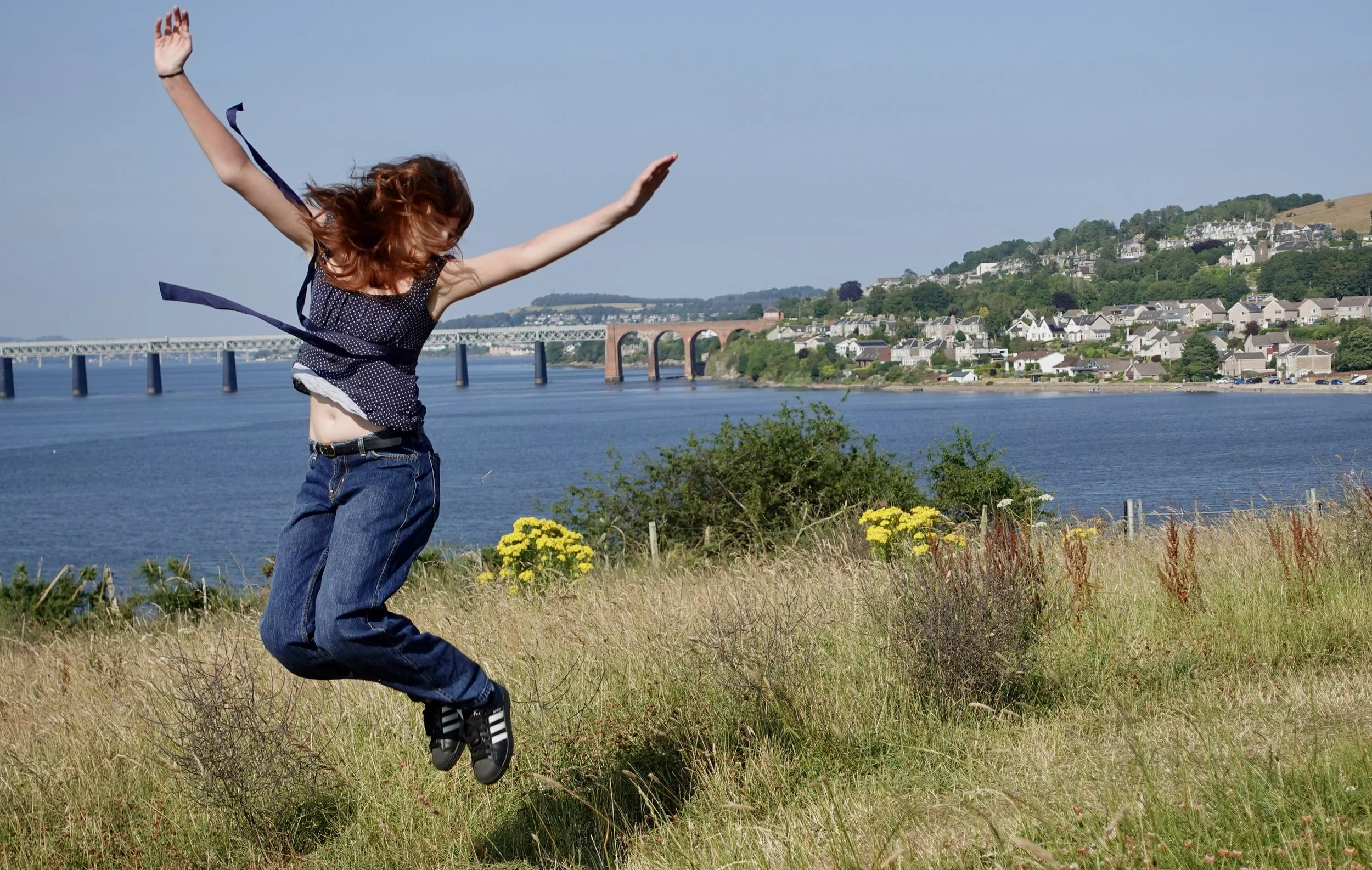 A person with red hair jumping in the air on a grassy field by the water, with a bridge and a hilly town in the background.