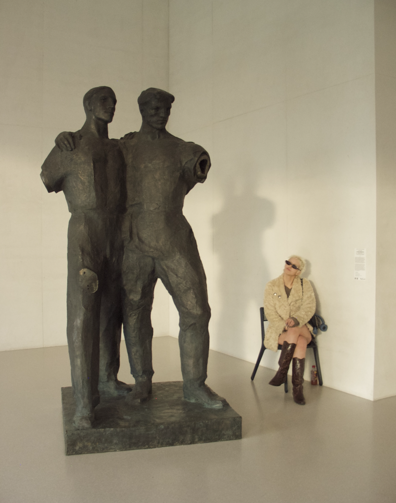 A woman sitting on a chair in front of a sculpture of two men. She is wearing sunglasses, a beige coat, boots, and has a purse on her lap. The sculpture depicts two male figures standing close together.