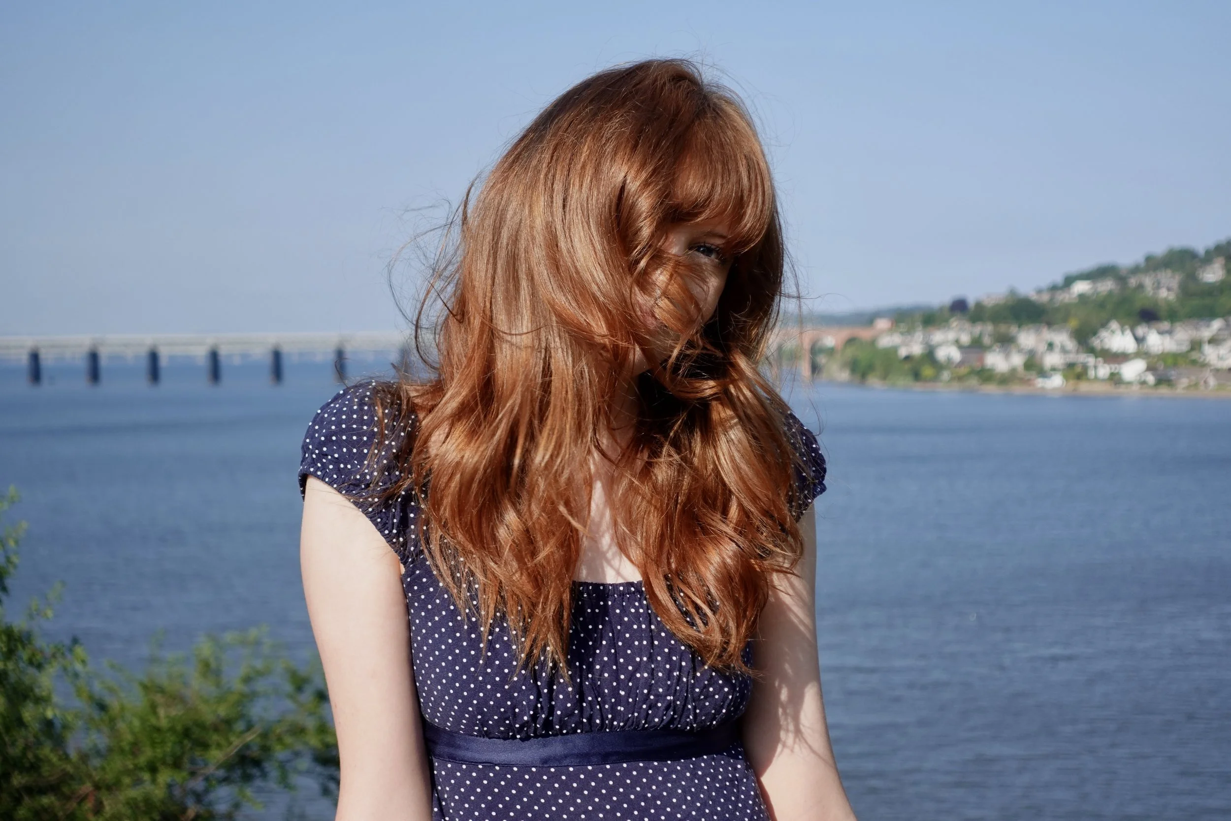 A young woman with long red hair smiling partially hidden behind her hair, wearing a navy blue dress with white polka dots, standing outdoors by a body of water with a bridge and hillside houses in the background.