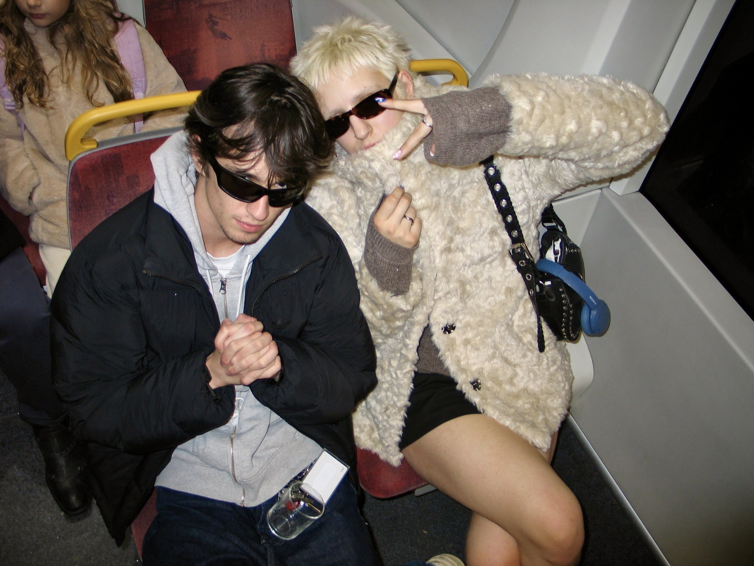 Two people sitting on a bus, both wearing sunglasses and holding hands. One person has long dark hair and the other has short blonde hair with spiky hair. They are sitting close to each other, with the person with blonde hair making a peace sign towards the camera. A third person can be seen in the background, and there are headphones hanging from the seat.