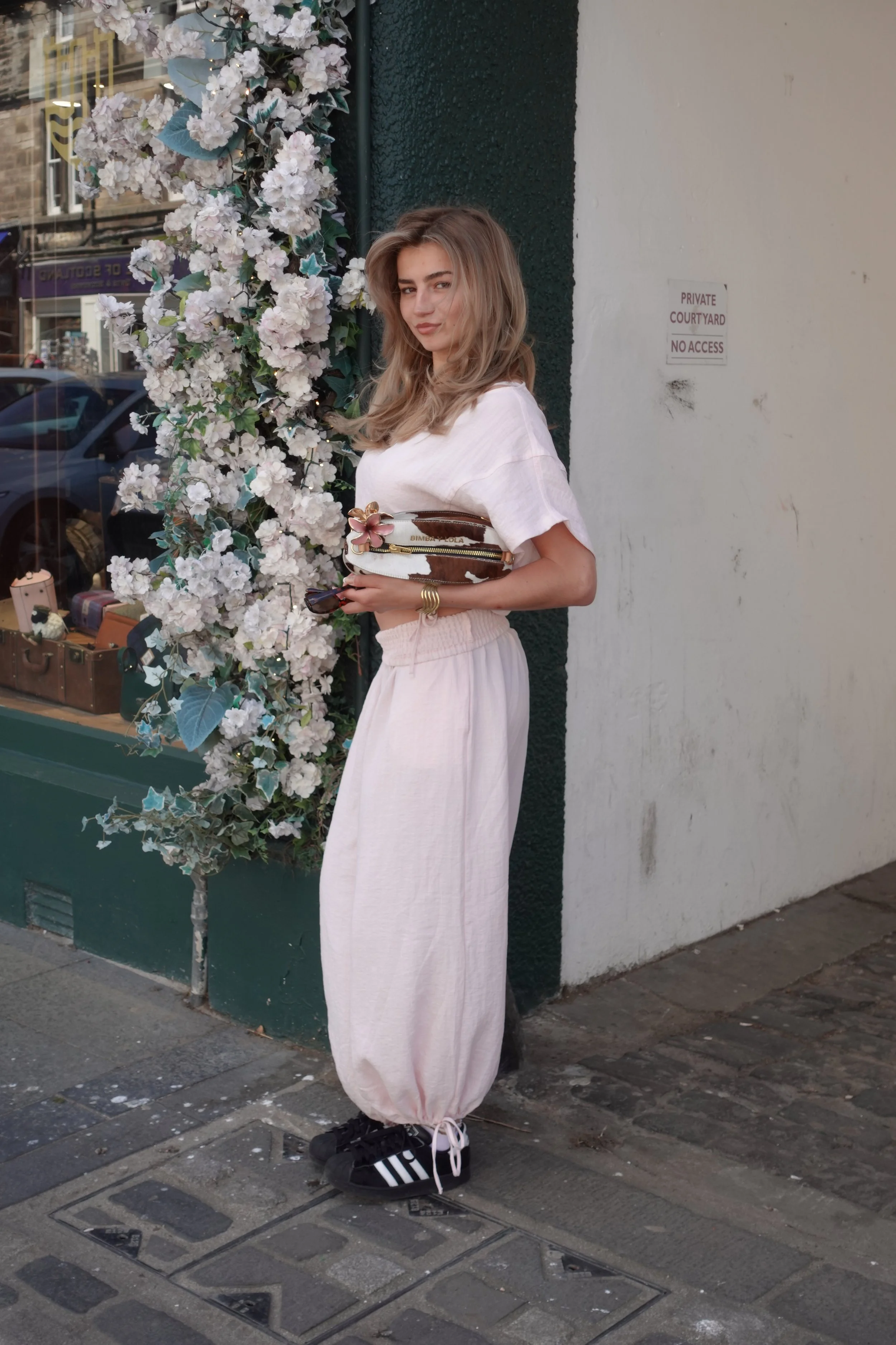 A young woman with light brown, wavy hair wearing a loose white top and cream-colored baggy pants holding a small crocodile skin handbag with a pink flower embellishment while standing on a sidewalk outside a store with a display of bags and souvenirs, next to a flower-covered wall, and a sign that reads 'Private Courtyard No Access' in the background.